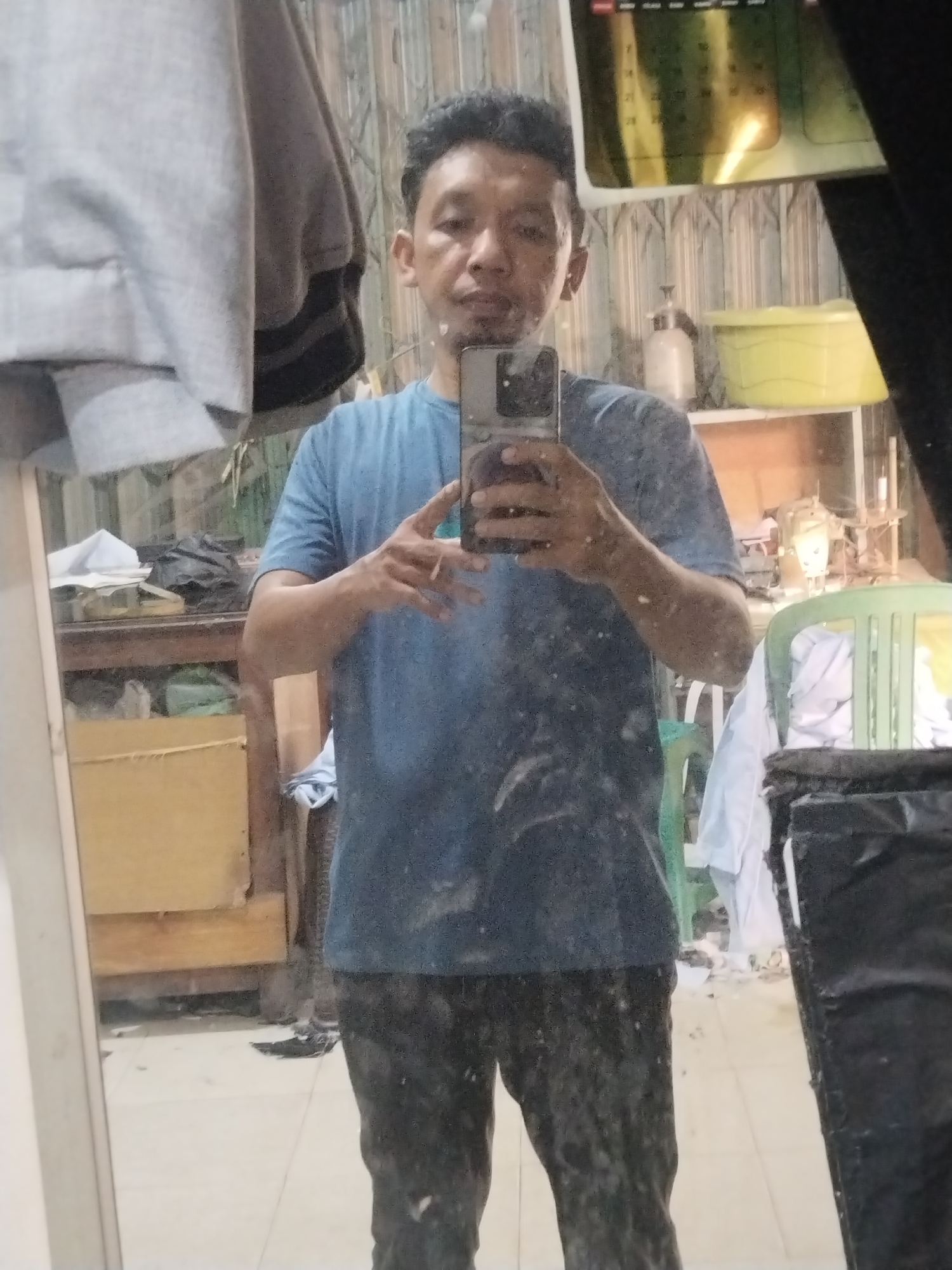 A full-body, front-facing mirror selfie of a man of Asian descent is captured in an indoor setting. The man is wearing a blue t-shirt and dark pants, and is holding a smartphone in his right hand, taking the picture. He has short, dark hair and appears to be looking at the phone's screen. The background is cluttered with various objects, including shelves with items on them, a yellow basin, a green chair, and what appears to be laundry. The wall behind him is covered in a corrugated metal material. The overall environment suggests a utilitarian or storage space. The image is taken in Unknown, Indonesia.