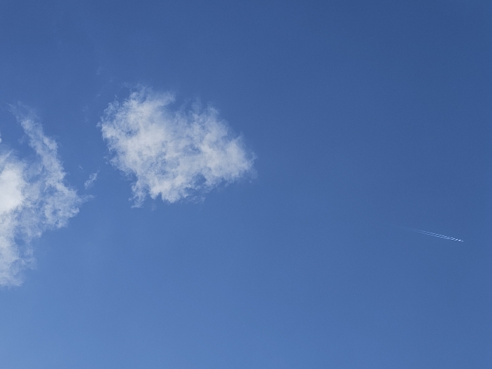 The scene depicts a clear blue sky above Donchevo, Bulgaria, under daylight conditions. Multiple white cumulus clouds are scattered across the upper half of the frame, with a larger cluster visible towards the upper right. A distinct, linear white contrail, characteristic of a high-altitude aircraft, originates from the lower-left corner and extends diagonally upwards and to the right, fading into the blue. No individuals, terrestrial objects, or specific interactions are visible within the frame, which focuses exclusively on the sky.