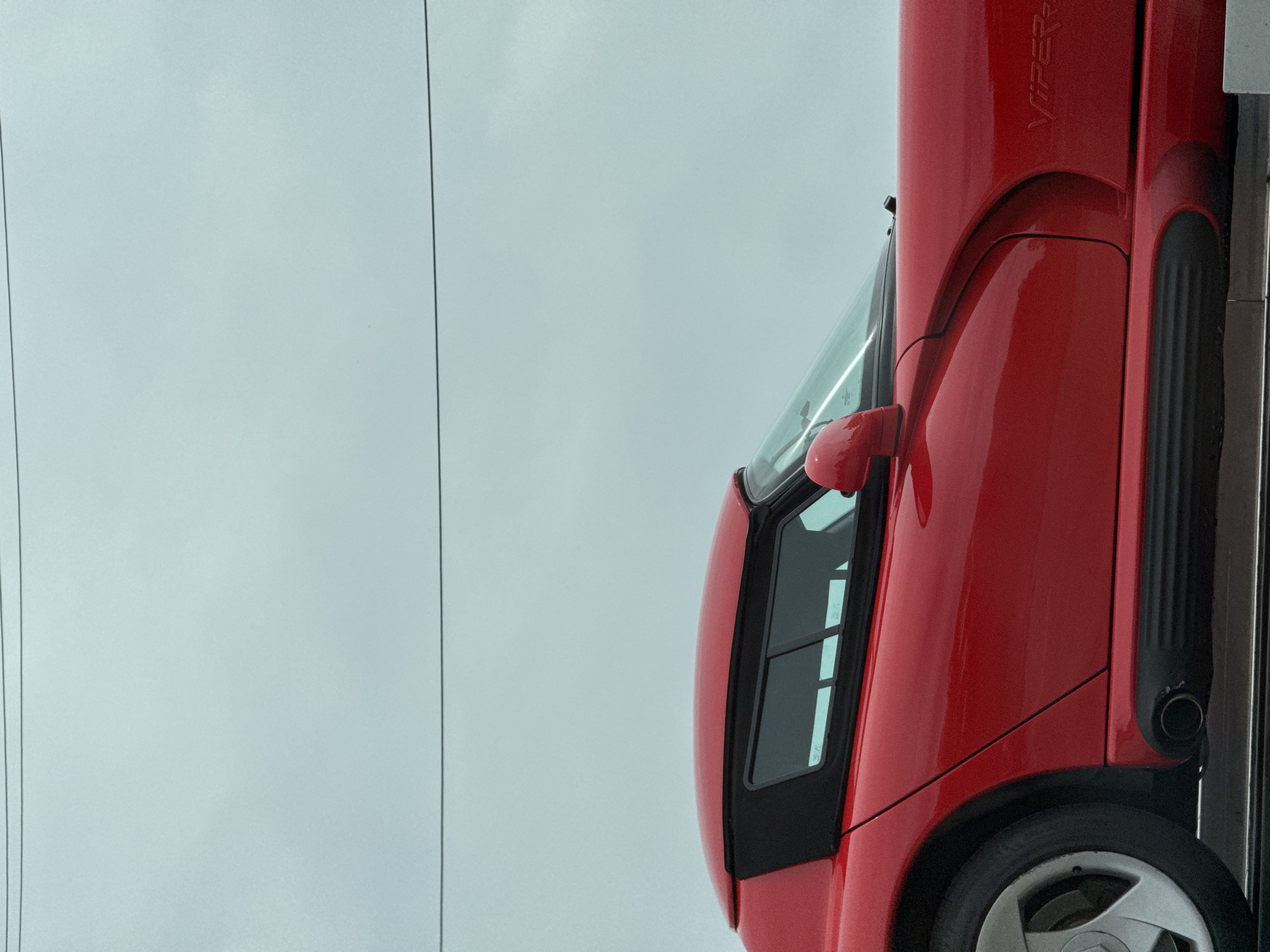 The image depicts the front right side of a red Dodge Viper RT/10. The car is positioned vertically, with the front facing upwards. The driver's side window, side mirror, wheel well, and part of the front tire are visible. The word "VIPER" is written in black on the red car. In the background, there is a cloudy sky, and two parallel power lines running vertically on the left side of the image. The exact location in Los Angeles cannot be determined from the image.