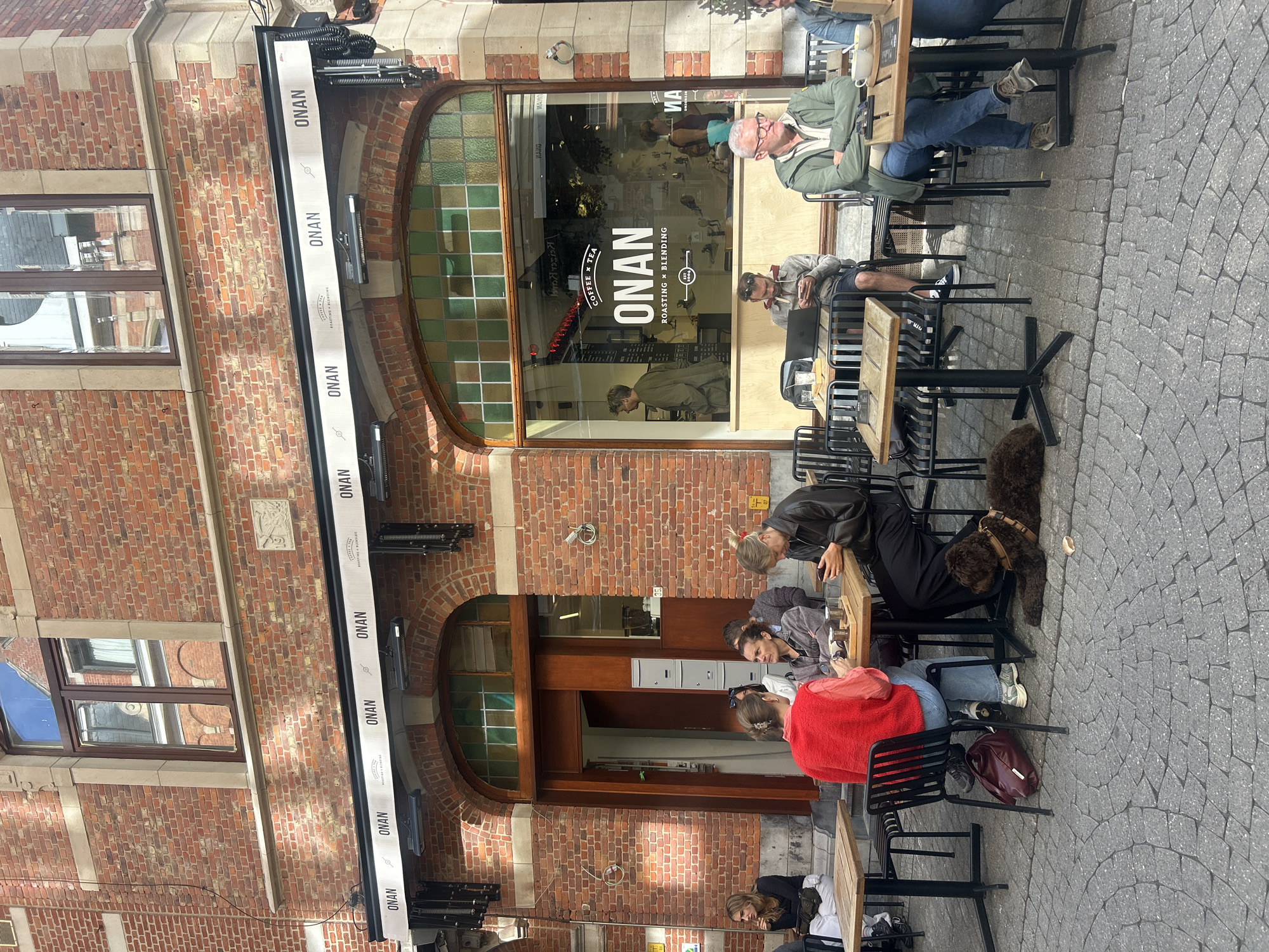 This is an exterior, eye-level, medium shot of a cafe, identified as ONAN Coffee & Tea, located on a cobblestone street in the city of Leuven, Belgium. The cafe features a brick facade with large arched windows and an awning displaying the ONAN logo. Several patrons are seated at tables outside the cafe. In the foreground, a person in a red vest and blue jeans sits at a table, interacting with another person across from them. To the right, a person in a black jacket and black pants is looking at their phone, with a brown dog lying on the ground next to them. Further to the right, a man in a green jacket and blue jeans is seated at a table, looking towards the right. Another man in a grey hoodie is seated at a table further back, looking at his phone and laptop. The overall scene depicts a casual outdoor dining or coffee setting.
