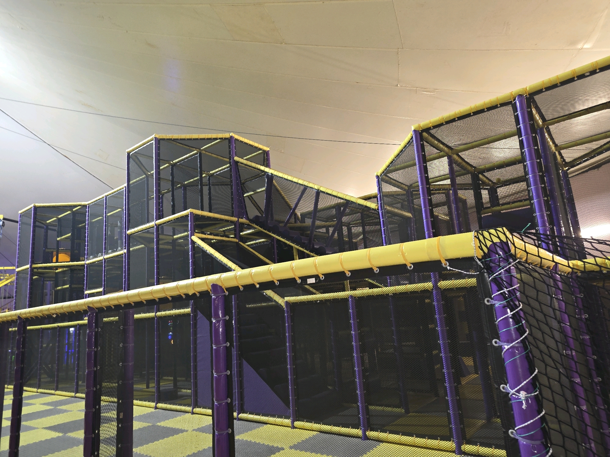 The image shows an indoor playground with purple and yellow structures. The playground features a multi-level maze-like design with netting and padded poles. The floor is covered in a yellow and grey checkered matting. The ceiling appears to be a taut fabric structure. No people are visible in the image. The scene is devoid of any context that would place it in Bodrum, Turkey, or any other specific location.