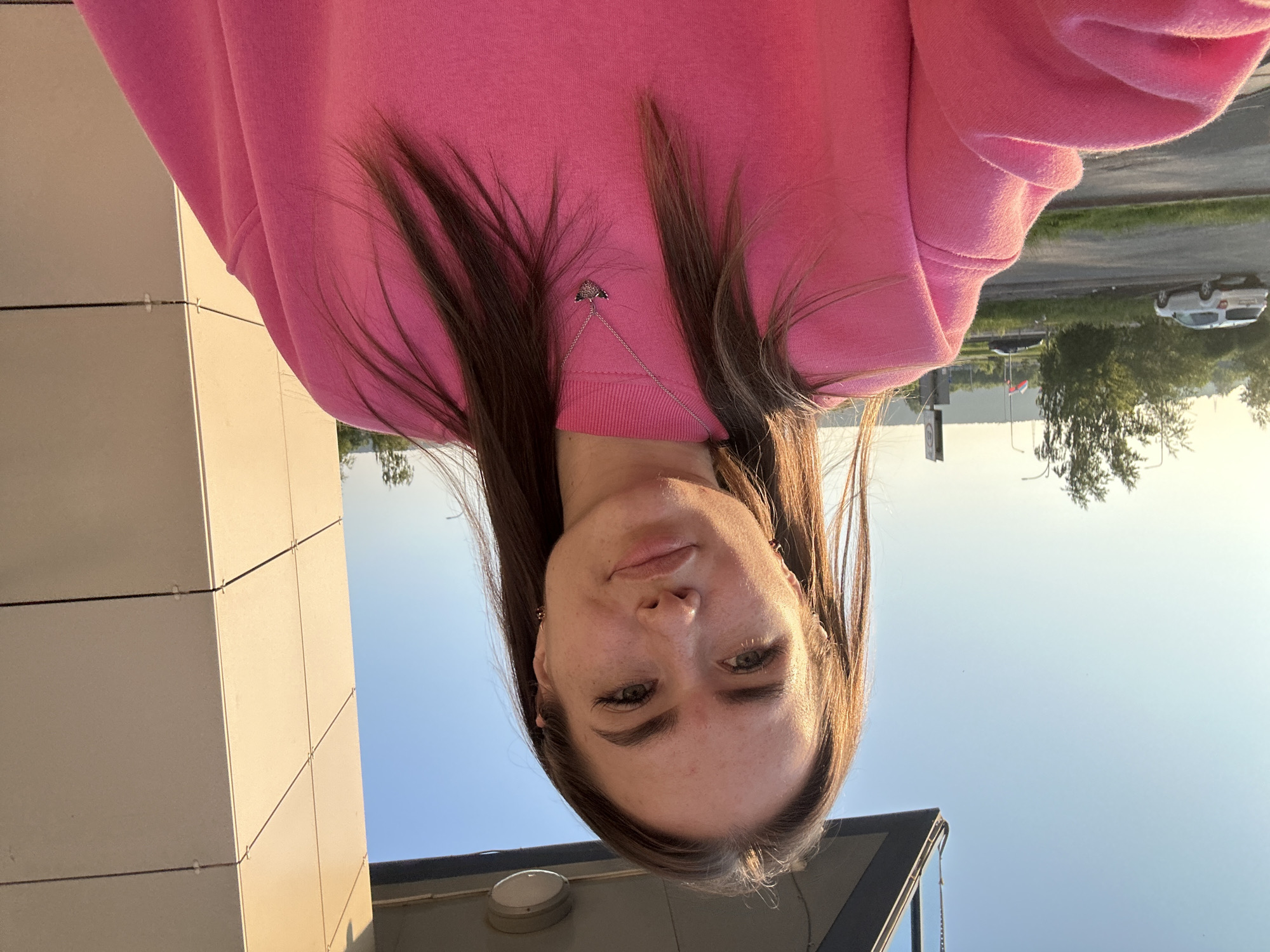A young woman with long, brown hair pulled back from her face, wearing a bright pink sweatshirt, smiles at the camera in a selfie. She has light skin, brown eyes, and her eyebrows are well-defined. She is wearing a delicate silver necklace with a small pendant. The background suggests an outdoor urban setting with a clear blue sky and a building with beige panels to her right. To her left, a street with a white car and trees is visible, with distant hills under a bright sky. The image appears to have been taken during golden hour.