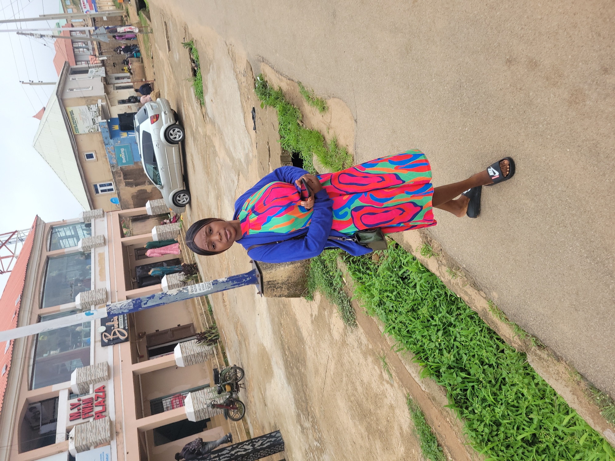 A woman stands on a paved road in Jos, Nigeria, holding a smartphone. She is wearing a blue cardigan over a colorful, patterned dress and black Nike slide sandals. Behind her, a building with "M.V. Iron Plaza" signage is visible, along with a parked motorcycle and a silver car. The environment appears to be a street with a drainage ditch filled with green grass and vegetation. Other buildings and individuals can be seen in the background.