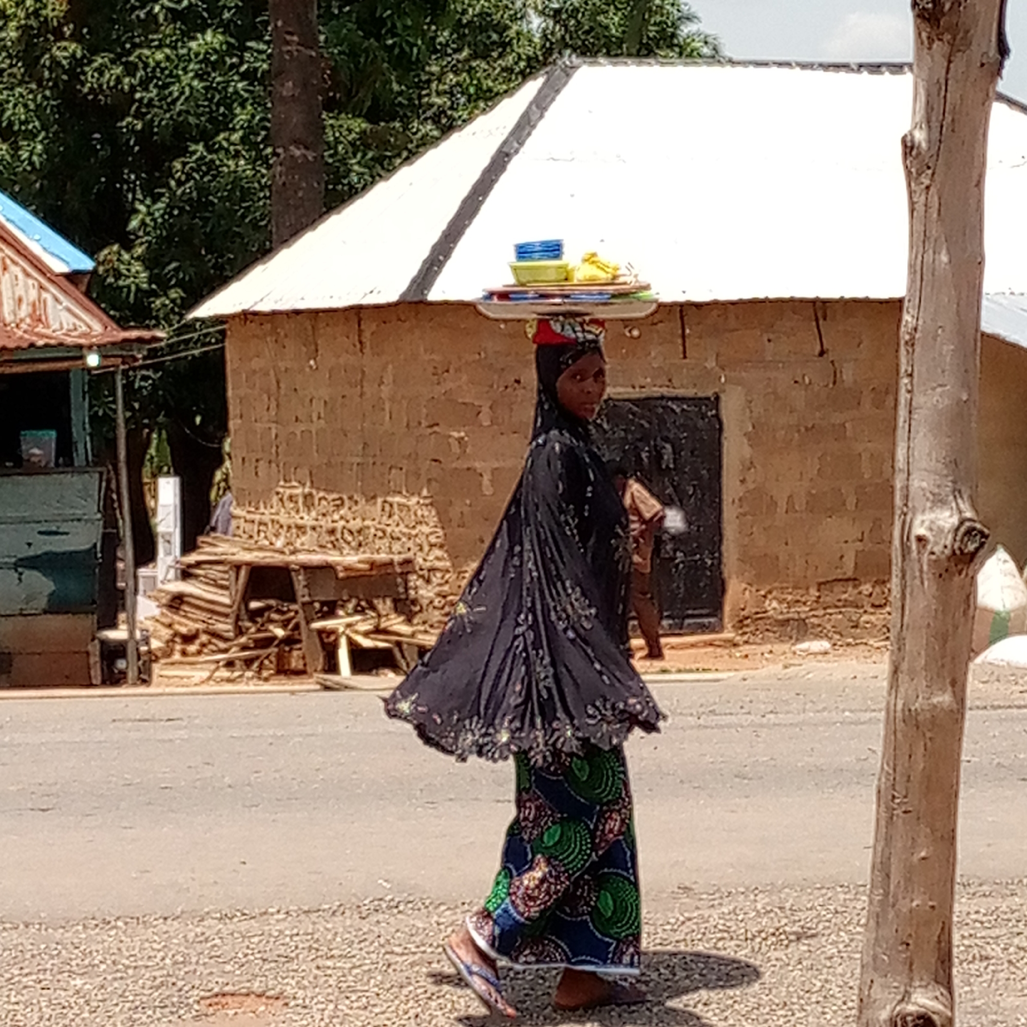 A woman is walking on a road in Mararaba, Nigeria. She has a tray of blue and yellow food items balanced on her head. She is wearing a black headscarf, a patterned black shawl, and a long, colorful patterned skirt. She is wearing flip-flops. In the background are buildings made of brick and corrugated metal. There is a pile of lumber behind her. A weathered tree trunk stands on the right side of the image. The road is asphalt, with a gravel shoulder. The sky is bright.