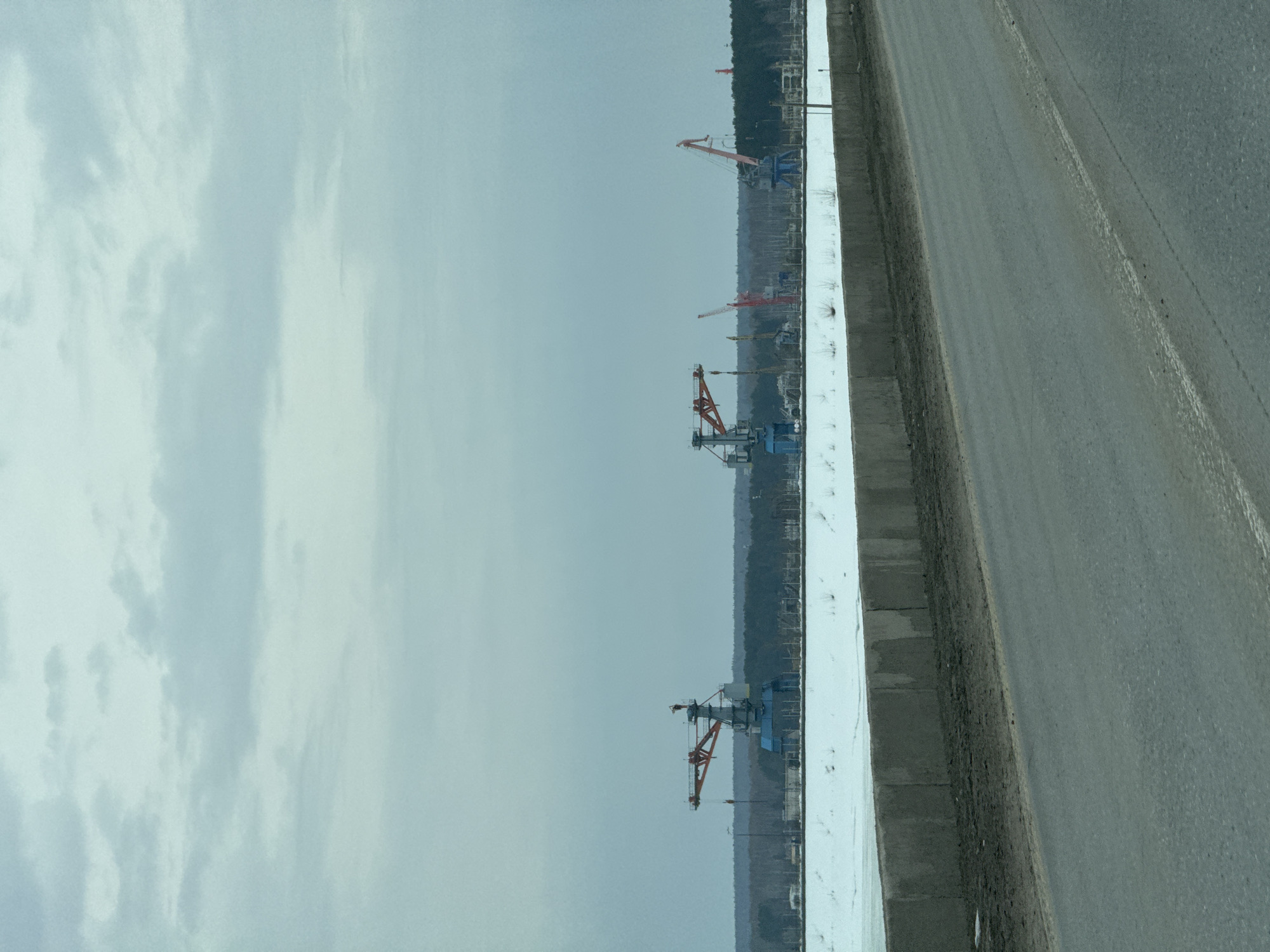 The image depicts a view from a vehicle, possibly a car, traveling along a road in what is assumed to be Chaikovsky, Russia, due to the presence of cranes. The road is bordered by a concrete barrier, to the left of which lies an industrial area with multiple cranes visible against a backdrop of trees. The sky is overcast. There are no people visible in the scene.