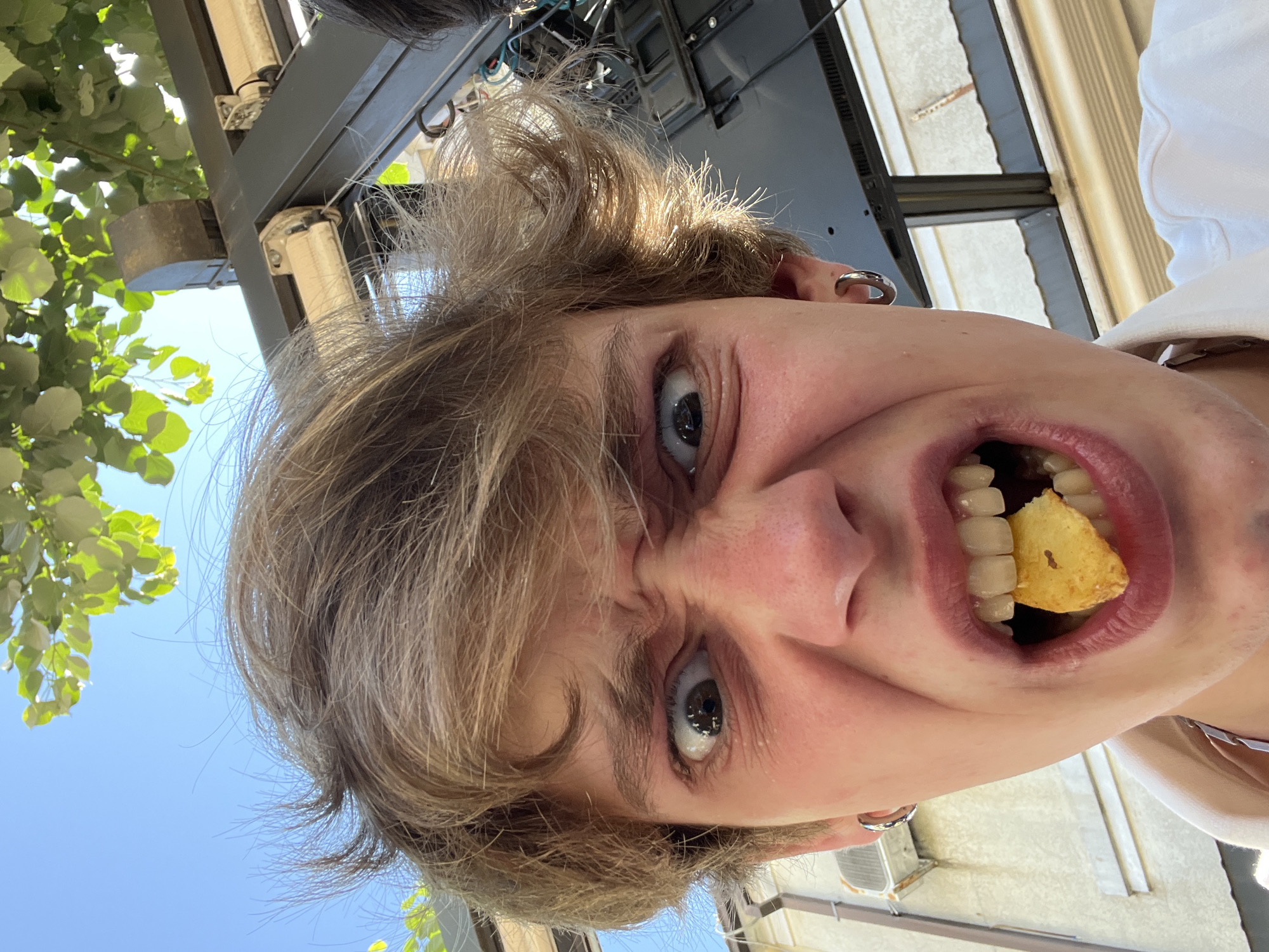A young person is captured in a close-up, low-angle selfie taken outdoors, possibly in Cetinje, Montenegro, on a sunny day. They have light brown, tousled hair and are wearing a white collared shirt. Their face is contorted in an exaggerated expression, with eyes wide open and eyebrows furrowed. A single potato chip is held between their teeth, and their mouth is open. Silver hoop earrings are visible in both ears.
In the background, lush green leaves from a tree are visible against a clear blue sky. A dark metal structure, possibly part of an outdoor seating area or awning, is positioned above and behind the person, with electrical wiring and components visible. The environment suggests an outdoor cafe or public space.