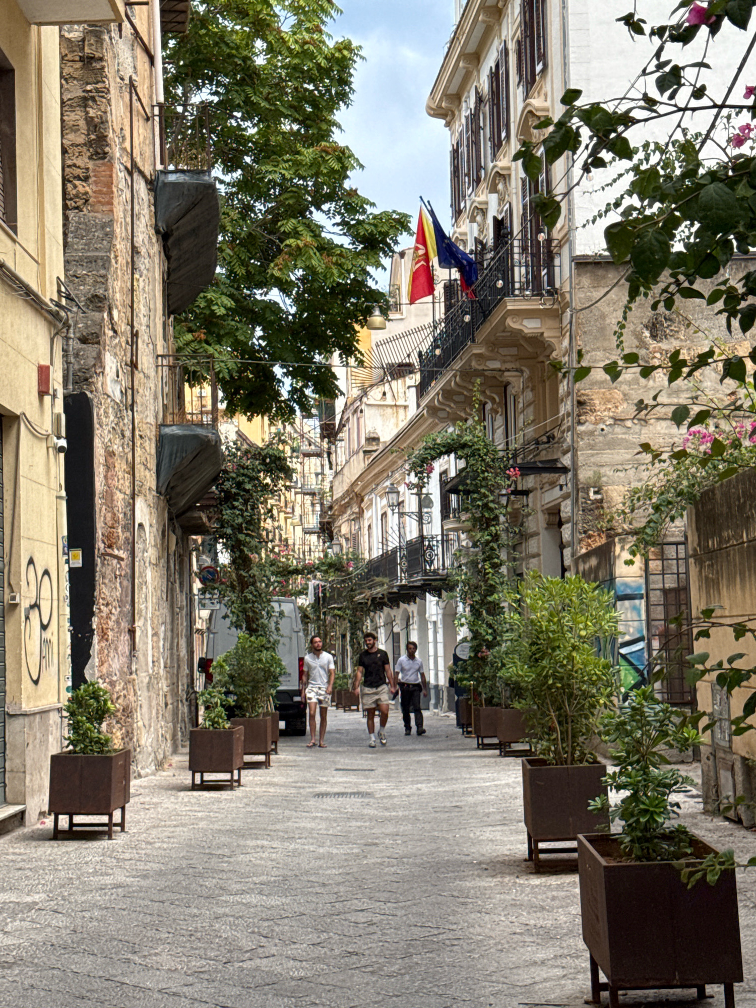 The image shows a narrow street in Palermo, Italy, likely a pedestrian walkway given the paving stones and lack of vehicular traffic. The architecture is characteristic of the city, with aged stone buildings lining both sides of the street, some featuring balconies with ornate railings. Several trees and plants are strategically placed in square planters, adding greenery to the urban environment. In the background, flags are visible hanging from a balcony, one of which appears to be the Italian flag. Three men are walking away from the camera down the street. The overall atmosphere is one of a sun-drenched Mediterranean street, with a sense of history and local charm.