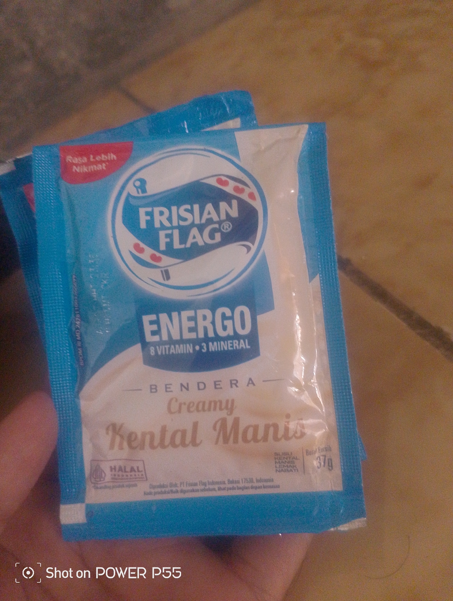 A hand holds two sachets of Frisian Flag Energo Creamy Sweetened Condensed Milk. The sachets are predominantly blue and white with red accents. The Frisian Flag logo is visible on the top sachet, along with the text "Rasa Lebih Nikmat" (More Delicious Taste). The second sachet displays "ENERGO 8 VITAMIN 3 MINERAL" and "BENDERA Creamy Kental Manis" (Sweetened Condensed Milk). A "HALAL" logo is present on the bottom left of the second sachet. The weight "37g" is indicated on the bottom right. The background is out of focus and appears to be a textured surface, possibly a floor or table. A watermark in the bottom left corner reads "Shot on POWER P55". The image has been taken from a close-up perspective, focusing on the product packaging. There is no discernible location in the city of Lebaksiu, Indonesia visible in the image.