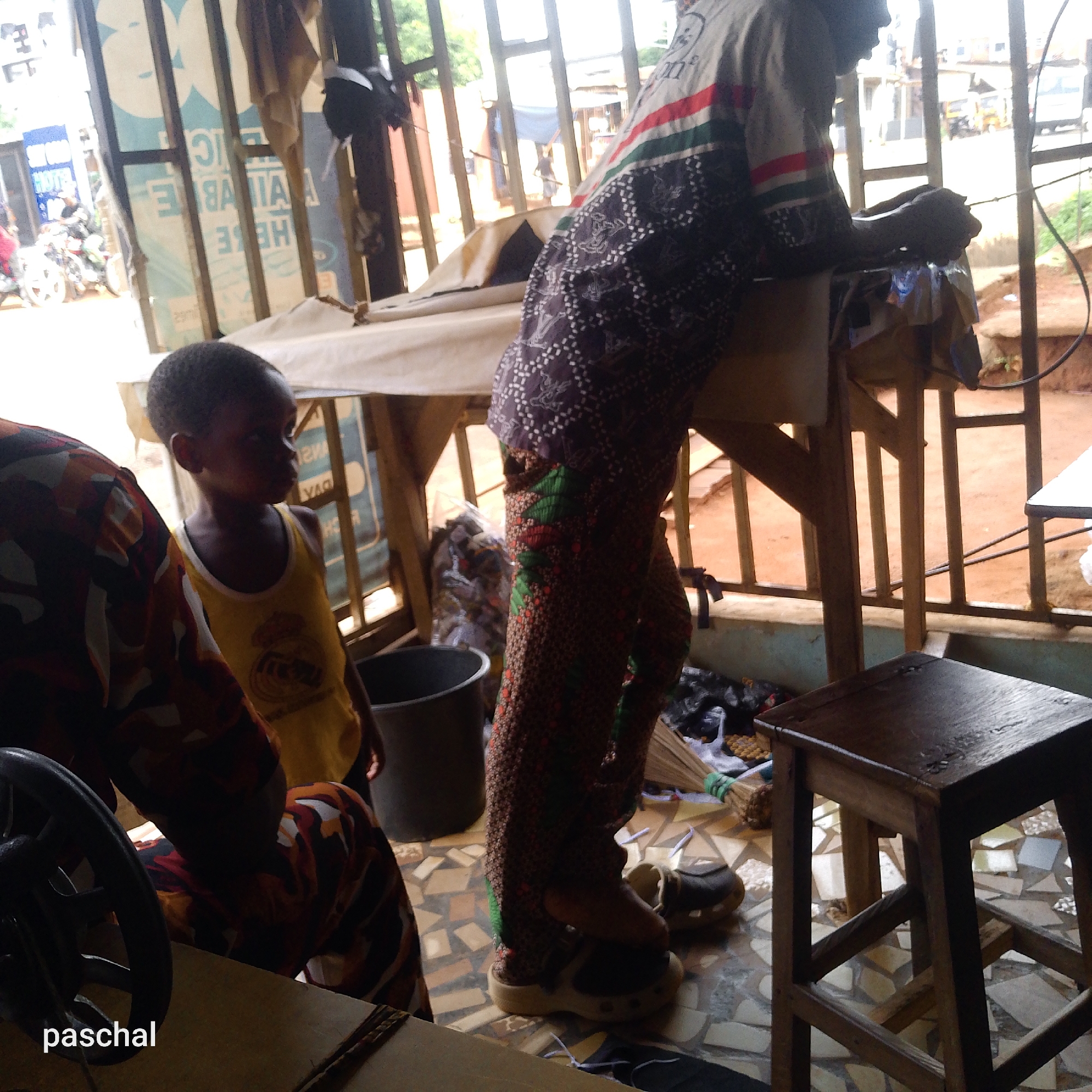 The image depicts a scene inside what appears to be a shop or workspace, possibly a tailor's shop. Inside, a child wearing a yellow shirt with a crest on it stands near an adult, presumably the tailor, who is leaning over a table. The tailor is wearing patterned clothing and sandals. A woman wearing patterned clothing is sitting near a sewing machine. A small wooden stool sits nearby. Outside the shop, there are glimpses of a street scene with other buildings, people, and vehicles. The location is in the city of Unknown, Unknown.