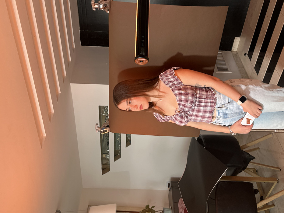 A young woman with brown hair and a plaid top stands against a brown backdrop in a well-lit room. She is wearing jeans and a smartwatch. A black studio light is visible above her, and various studio equipment is scattered around. The room features modern design elements, including linear wall decor and a black and white striped area.