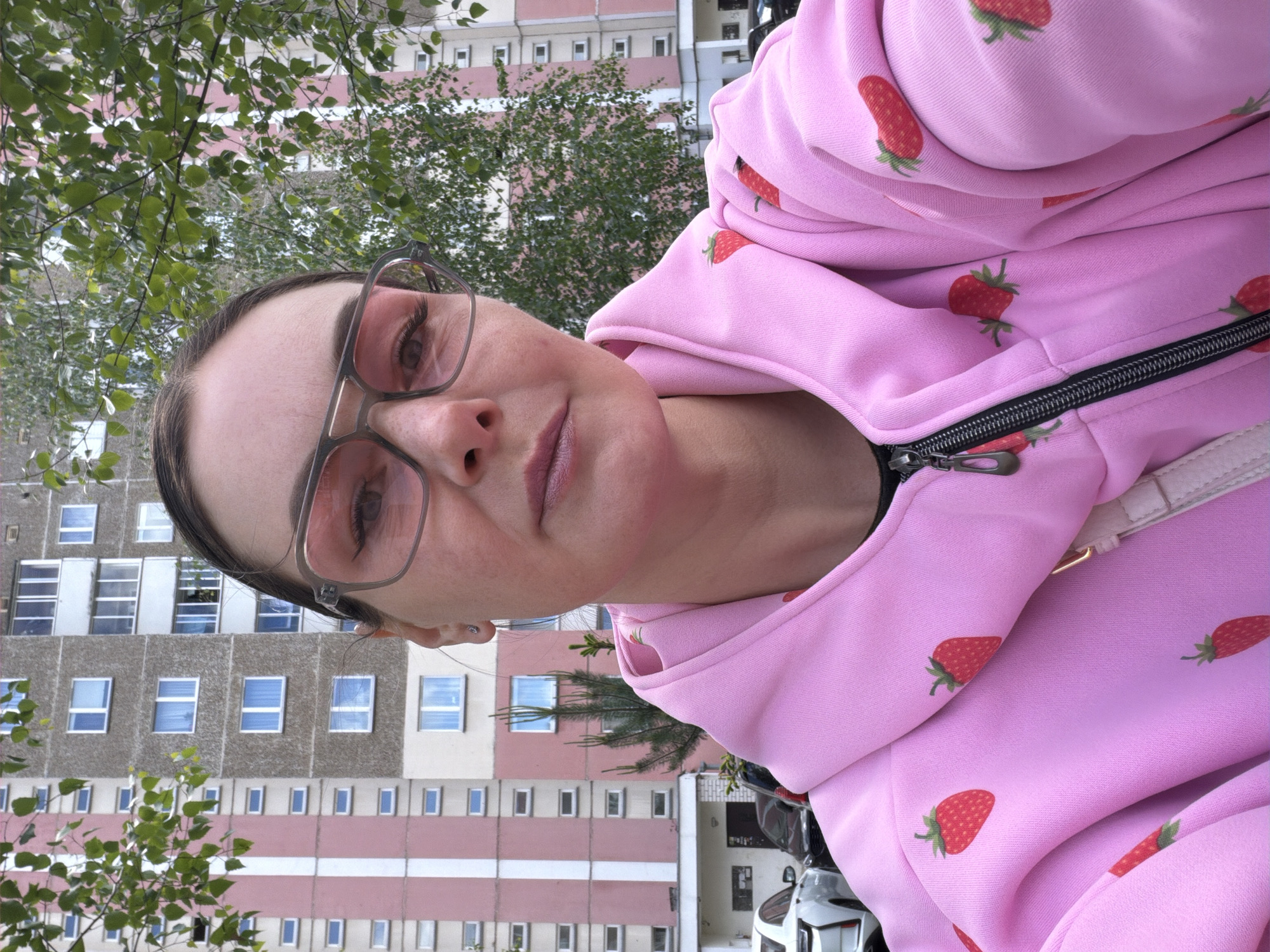 A woman with glasses and a pink strawberry-patterned hoodie poses, with an apartment building and trees in the background. The building has multiple rows of windows and a pink and gray facade. Parked cars are visible near the base of the building. The exact location in the city of Unknown, Unknown, is not determinable.