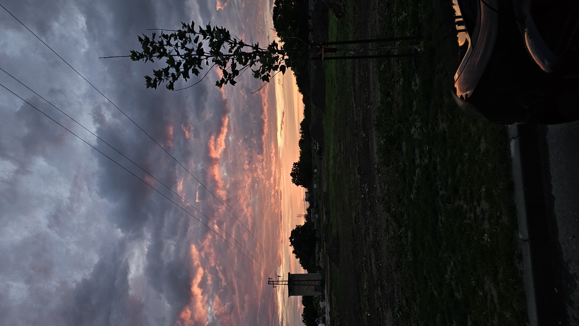 This image captures a twilight scene with a dramatic sky painted with hues of pink, orange, and purple, breaking through a blanket of dark, heavy clouds. In the foreground, the silhouette of a car is visible on the right, its glossy surface reflecting the vibrant sky. Beyond the car, a patch of dark grass and a curb lead to a muddy, uneven ground scattered with piles of dirt, suggesting a construction site.
Further back, a solitary young tree stands to the right, its dark leaves stark against the colorful sky. In the middle ground, a rectangular concrete structure with a metal tower on top is prominent on the left. Several trees form a dark line in the distance, and more indistinct structures and mounds of earth are visible across the landscape. Four power lines stretch diagonally across the sky, adding a linear element to the composition. The overall impression is of an industrial or developing area at dusk, with nature's dramatic display overhead.
The scene is identifiable as being near the area of the construction site for the Plovdiv bypass, close to the junction with the main road connecting Plovdiv to its southwestern suburbs. This location is on the outskirts of Plovdiv, Bulgaria.