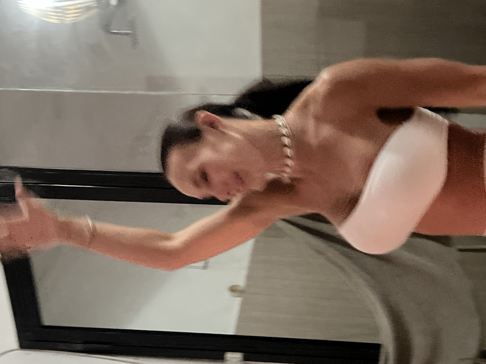 In an indoor setting, a blurred individual is captured in motion, raising their right arm. They are wearing a white bandeau top and a shell necklace. The background features a large mirror, a doorway, and what appears to be a tiled wall. The lighting suggests an artificial light source, possibly a lamp visible in the mirror's reflection. The overall image is unfocused, giving a sense of movement or a candid snapshot. The location is not specified in the image itself, nor can it be determined from the visual information.