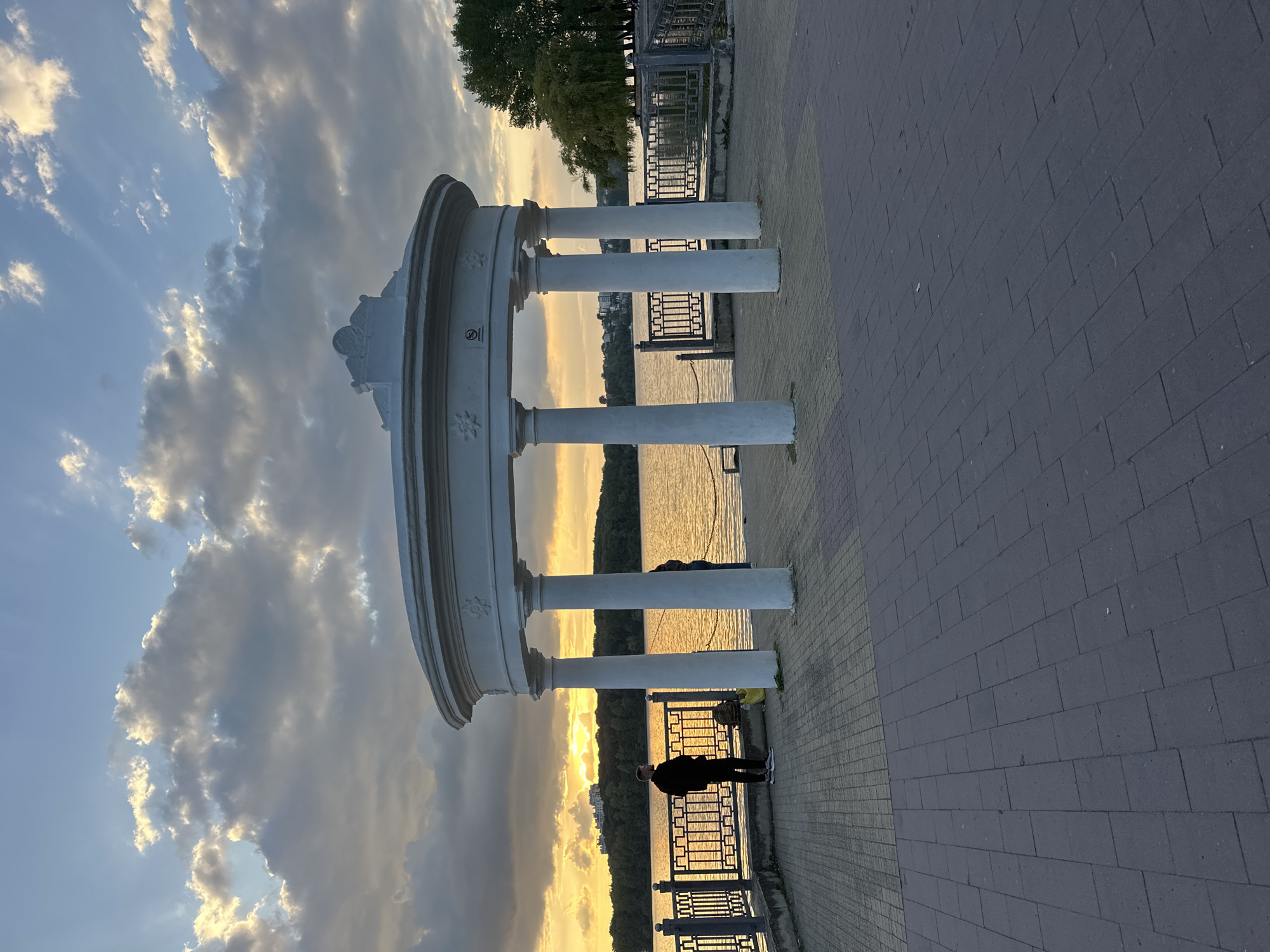 The image depicts a waterfront scene in Ternopil, Ukraine, likely at sunset. A white, classical-style rotunda with eight columns stands prominently in the foreground, framing a view of a calm body of water. The sky above is filled with dramatic clouds, illuminated by the warm, golden light of the setting sun, which casts a glow on the water's surface.
On the paved promenade to the left of the rotunda, a person dressed in dark clothing stands facing the water, their back to the camera. Another person, also in dark clothing, is partially visible within the rotunda. A decorative metal railing runs along the edge of the waterfront. In the distance, across the water, a tree-lined shore with buildings is visible. The promenade itself is paved with rectangular stones.