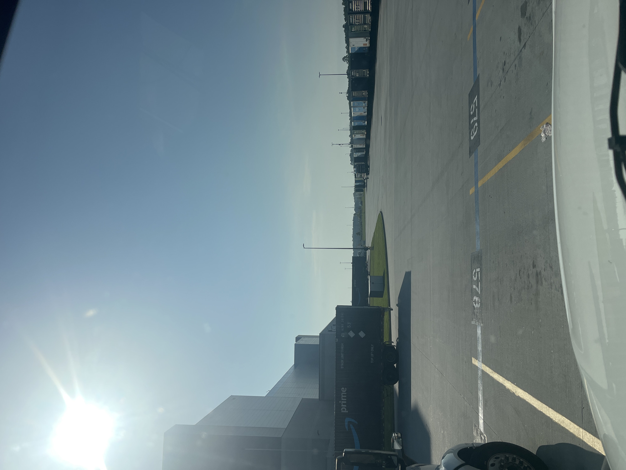 The image depicts an outdoor scene at what appears to be a logistics or distribution center. The sun is shining brightly, suggesting it is daytime, possibly morning or afternoon, with a clear blue sky. The setting is an asphalt lot marked with yellow and blue lines, indicating parking or loading bays.
Prominently featured are several large shipping containers, many bearing the Amazon Prime logo and branding. Some containers are stacked, while others are on the ground. One visible container has text that reads "STACK: UNIT 3 HIGH" and "TOP LIFT ONLY". Another container has "prime" and a blue Amazon logo. There are also what appear to be semi-trucks or tractor-trailers parked in a line. Some of these vehicles are white, and one in the foreground is partially visible and appears to be white as well.
In the background, a large, modern building with a reflective facade is visible, likely the distribution center itself. The lot is also lined with light poles. A patch of green grass with some bushes is visible in the middle ground. The ground is concrete or asphalt, with some scattered debris and water marks. There are numbers painted on the ground, such as "578" and "579", likely identifying parking spots or bays. The overall impression is one of a busy industrial area focused on shipping and logistics. No people are visible in the image.