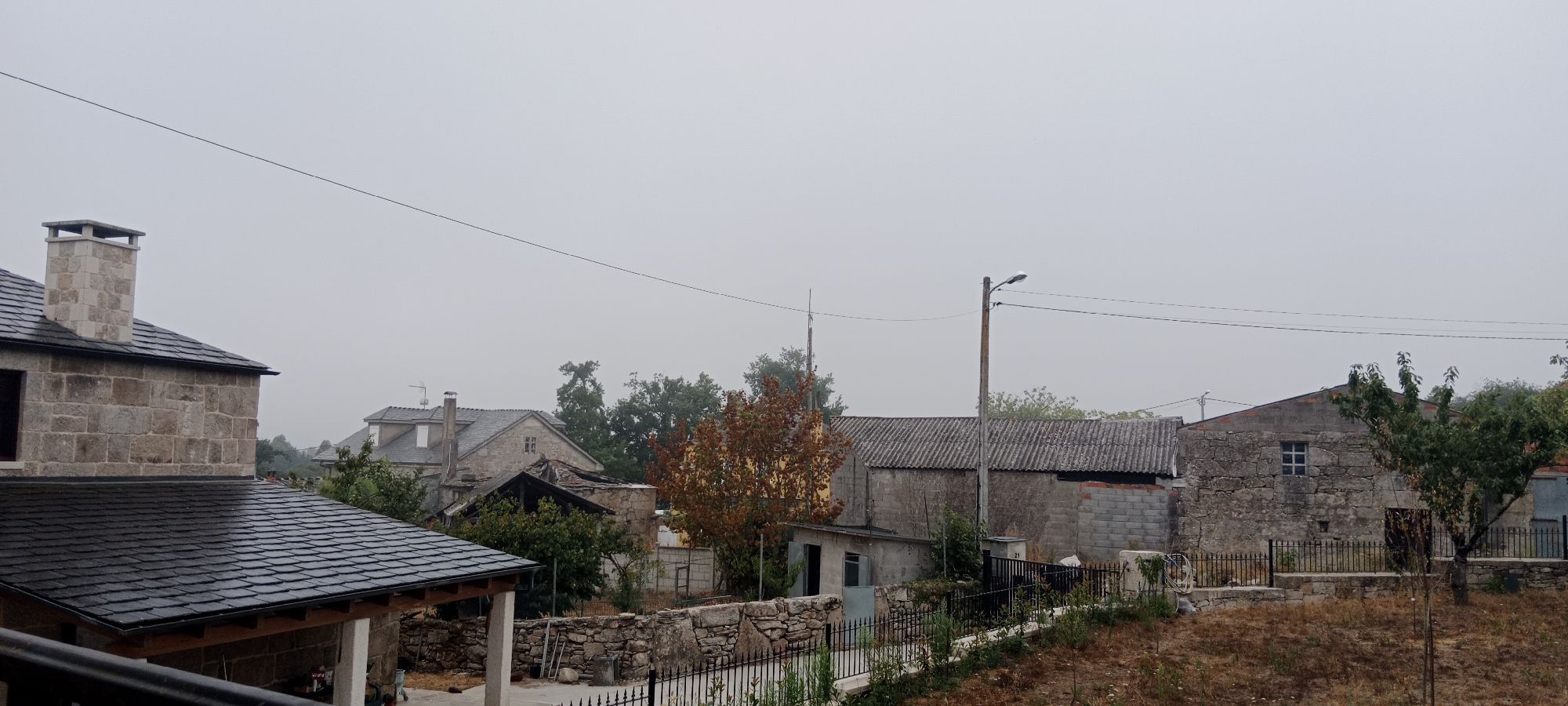 In Adai, Spain, on a cloudy day, a rural village scene unfolds. The foreground features the dark slate roof of a building, with a stone chimney on an adjacent stone house. Beyond this, a cluster of stone and brick houses are visible, some with tiled or corrugated roofs. Trees with sparse foliage, some bearing red-brown leaves, dot the landscape. A street lamp stands prominently in the middle distance. A stone wall, with metal fencing, encloses some of the properties. The sky is overcast and a thin electrical wire stretches across the frame.