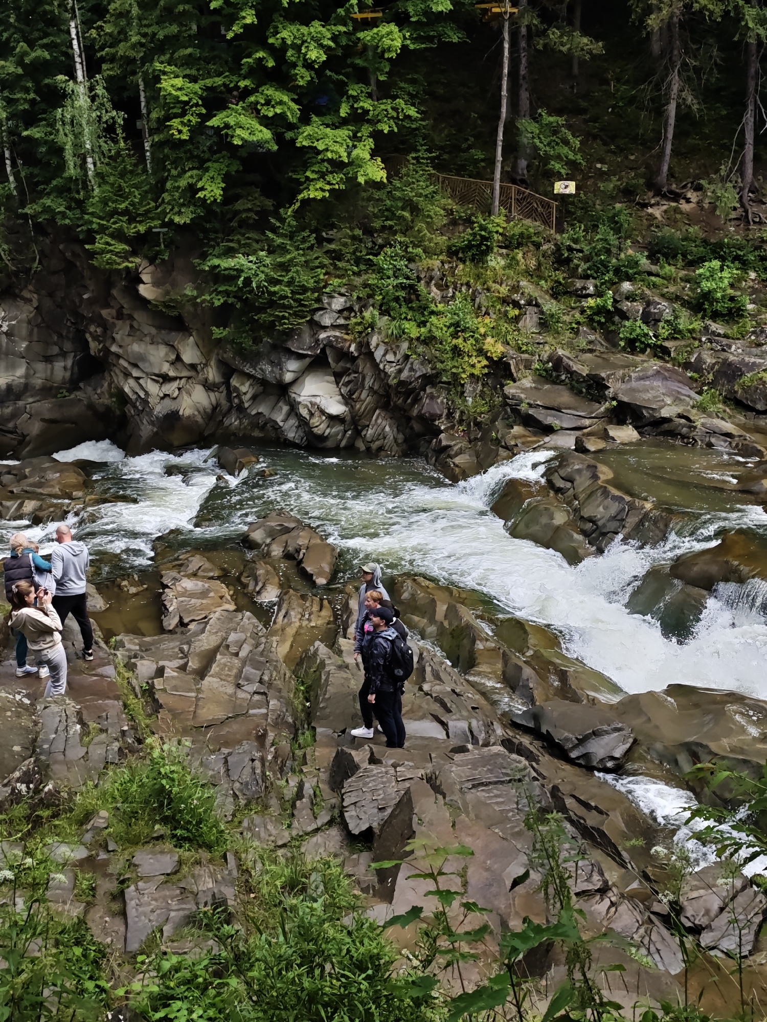 A group of tourists are standing on the rocky bank of a powerful mountain river in Yaremche, Ukraine. The river flows rapidly over numerous large, smooth stones, creating white water rapids and cascades. Lush green trees and vegetation cover the steep, rocky slopes surrounding the river. The tourists, dressed in casual clothing and backpacks, appear to be observing the natural scenery, with some taking photographs. The overall impression is one of a scenic natural attraction within a mountainous region.