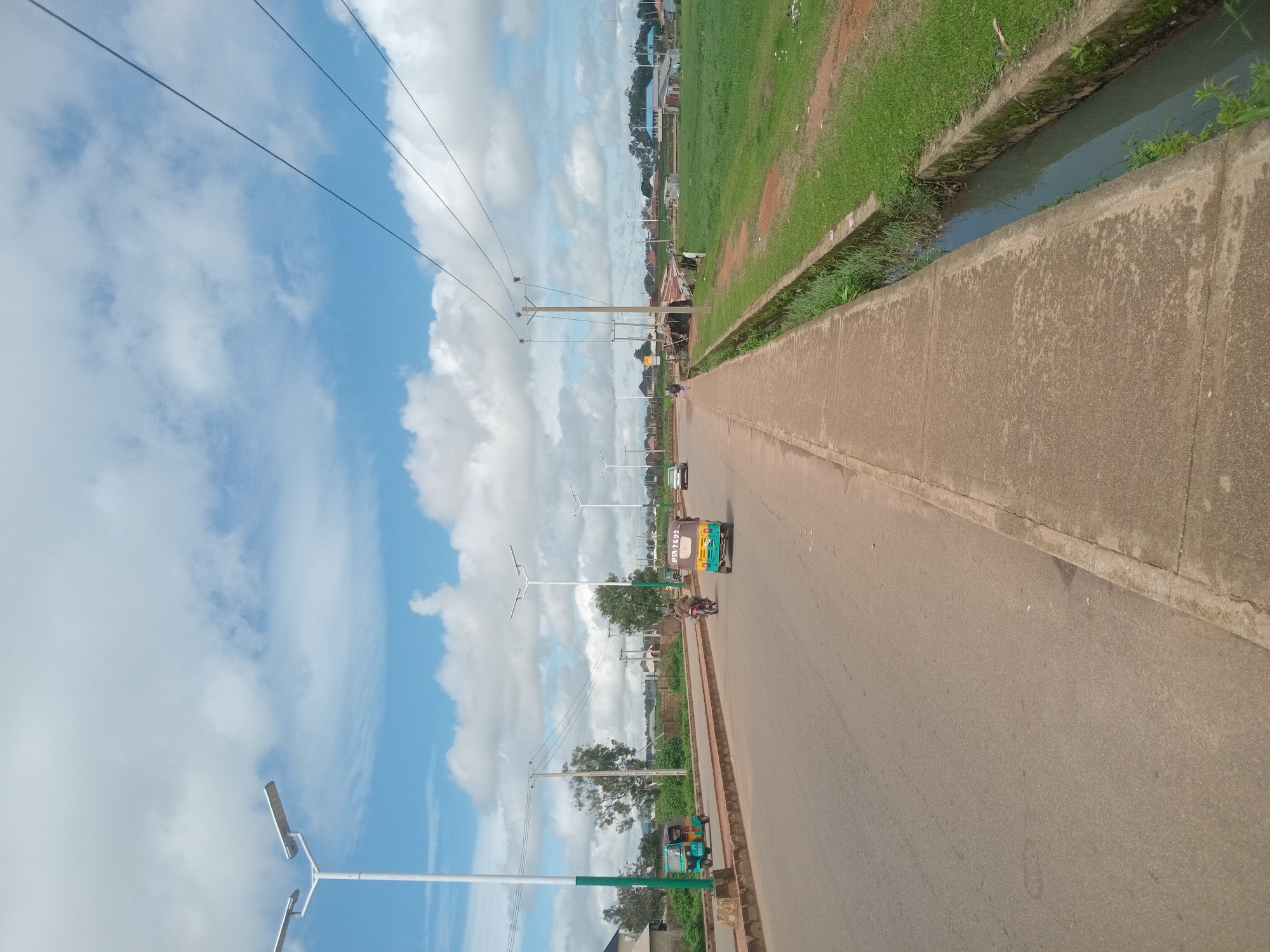 In Bukuru, Nigeria, a wide paved road stretches through a developing urban area under a partly cloudy sky. The road is bordered on the left by a raised median with sparse vegetation and a sidewalk. On the right, a concrete drainage ditch runs alongside a grassy area. Several modern streetlights with solar panels are visible along the median. Traffic includes a green and yellow tricycle, a motorcycle with a rider, and a silver car moving away from the viewer on the road. A few buildings and structures, including what appears to be a makeshift stall, are situated in the background, interspersed with trees and more greenery. Power lines are strung across poles on the right side of the road.