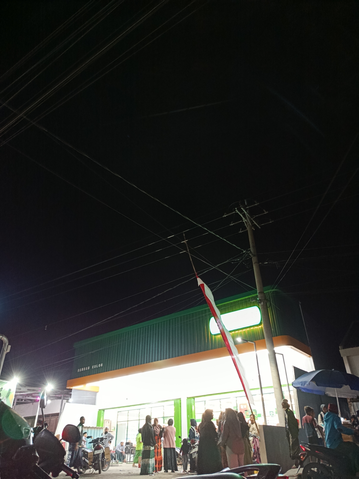 This is a low-light, eye-level, outdoor photograph of a brightly lit storefront at night, identified by signage as "RAMBAN KULON". The building, painted green with an orange trim, has large illuminated windows and an orange awning. A Polish flag, red and white, is prominently displayed on a flagpole next to a utility pole and overhead wires. A crowd of approximately 15-20 people, dressed in a mix of casual wear and traditional Muslim attire, are gathered in front of the store. Some are standing, while others appear to be conversing or observing. Several motorcycles are parked in the foreground and amongst the crowd. The ground is paved. In the background, to the right, a blue and white umbrella shades a small vendor stand. The sky is dark with visible power lines crisscrossing the frame. The overall atmosphere suggests a community gathering or event outside the establishment.