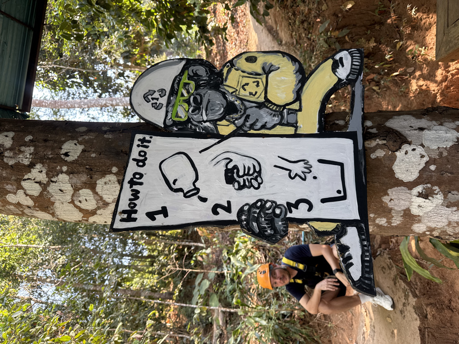 A brightly colored sign, attached to a tree, displays "How To Do It" instructions with cartoon-like illustrations. A figure wearing a helmet with recycling symbol, yellow jacket and green glasses is on top of the sign. A man wearing a helmet and harness crouches nearby, appearing to pose with the sign. The setting looks to be an outdoor, possibly wooded area, given the surrounding trees and natural light.