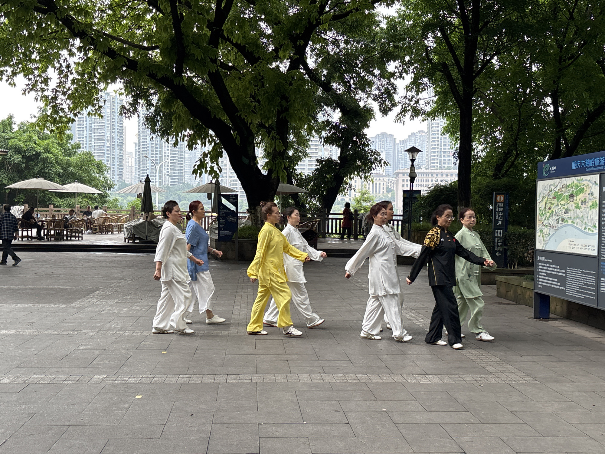 A group of seven women are walking on a paved walkway in Yuzhong District, China. They are practicing Tai Chi, dressed in matching silk-like outfits in various colors (white, light blue, yellow, black, and light green). The walkway is bordered by trees and foliage. In the background, there is a cityscape of tall buildings and a seating area with tables and umbrellas. A tourist information sign with Chinese writing is visible on the right.