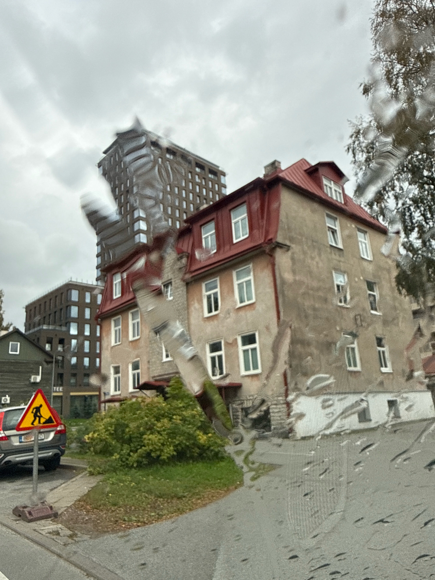 The image shows a view through a wet window of buildings in Tallinn, Estonia. In the foreground, on the left, is a grey car parked next to a yellow triangular construction warning sign depicting a person with a shovel. Behind the car is a low, dark wooden building with a white square window. To the right of this, and slightly further back, is a multi-story apartment building with a red pitched roof and beige walls, featuring numerous white-framed windows. Behind this building, and taller, is a modern building with a dark facade and patterned windows. The sky is overcast, and the weather appears to be rainy, as indicated by the water droplets on the window in the foreground. The overall impression is of an urban street scene on a cloudy day.