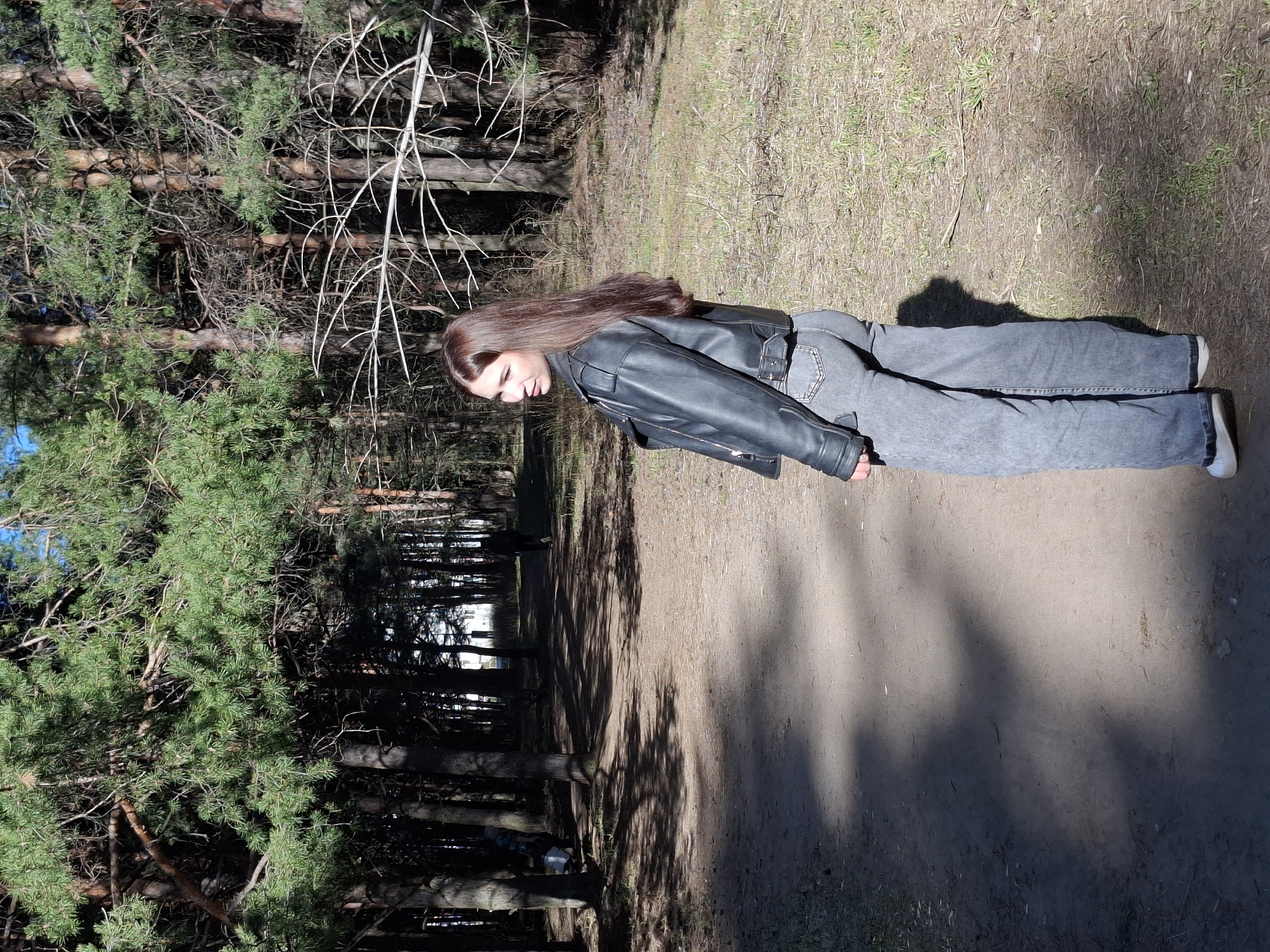 The image depicts a woman standing on a dirt path in a wooded area. She has long brown hair and is wearing a black leather jacket, gray jeans, and white shoes. The woman appears to be looking down. The surrounding environment consists of trees, including pine trees with visible bark and green needles, and a dirt path. Shadows cast by the trees are visible on the path. The location is suspected to be in Chaikovsky, Russia, given the presence of pine trees, a common feature of forests in that region.