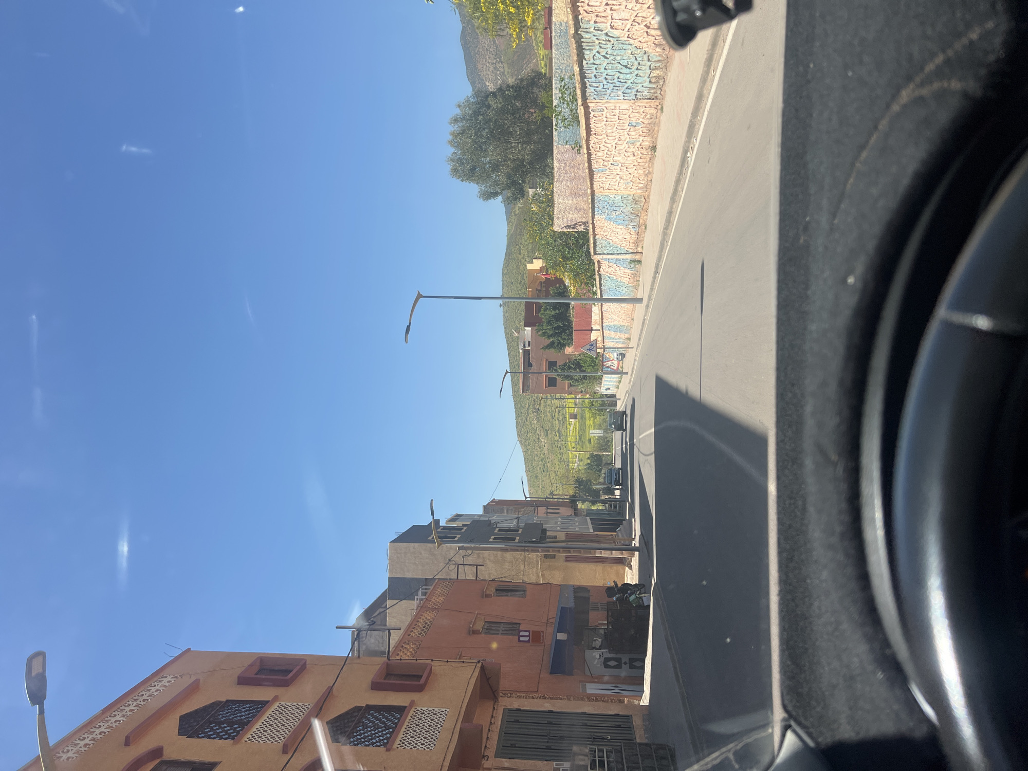 The image shows a street in Ouaouizert, Morocco. The view is taken from inside a vehicle, with the car interior visible on the right side. The street is paved and lined with buildings of varying heights and colors, primarily in shades of orange and brown. Some buildings have detailed window designs. Streetlights are present along the road. In the distance, a hill or mountain covered with green vegetation is visible under a clear, blue sky. A painted wall runs along the right side of the road, adding a colorful accent to the scene. The overall impression is that of a sunny day in a small town.