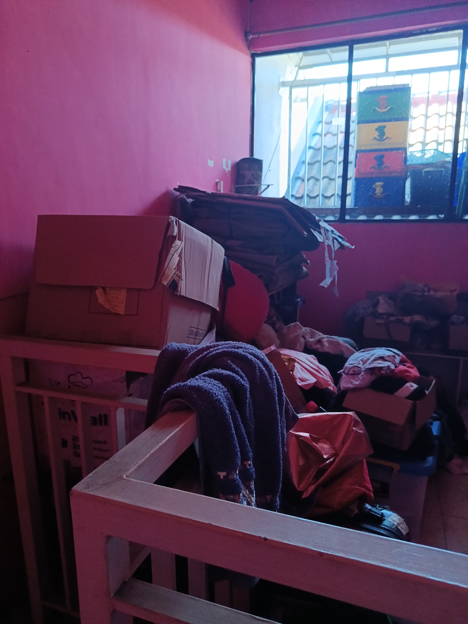 The image depicts a cluttered room, likely a storage area or a less-used living space, in Keboananom, Indonesia. The walls are painted a vibrant pink. A window with bars looks out onto tiled rooftops and what appears to be a fence or trellis. Dominating the foreground is a wooden structure, possibly a railing or part of a piece of furniture, draped with a dark blue knitted item.
To the left, several cardboard boxes are stacked. The largest is a light brown box with flaps partially open, revealing tape and some yellowing labeling. Another box, slightly smaller and also brown, is positioned behind it, showing a label with "530x420x450".
Towards the right and in the background, the scene becomes more chaotic. A pile of dark fabric, possibly blankets or clothing, is stacked high. A bright red, rounded object, perhaps a cushion or a deflated ball, is visible. Various other boxes and containers are present, some open and filled with what appears to be clothing and other soft items in shades of pink, white, and red. A blue plastic bin is also visible. A bright red plastic bag, partially filled, sits on top of a black object, possibly a shoe with a label. The overall impression is one of disarray and storage.