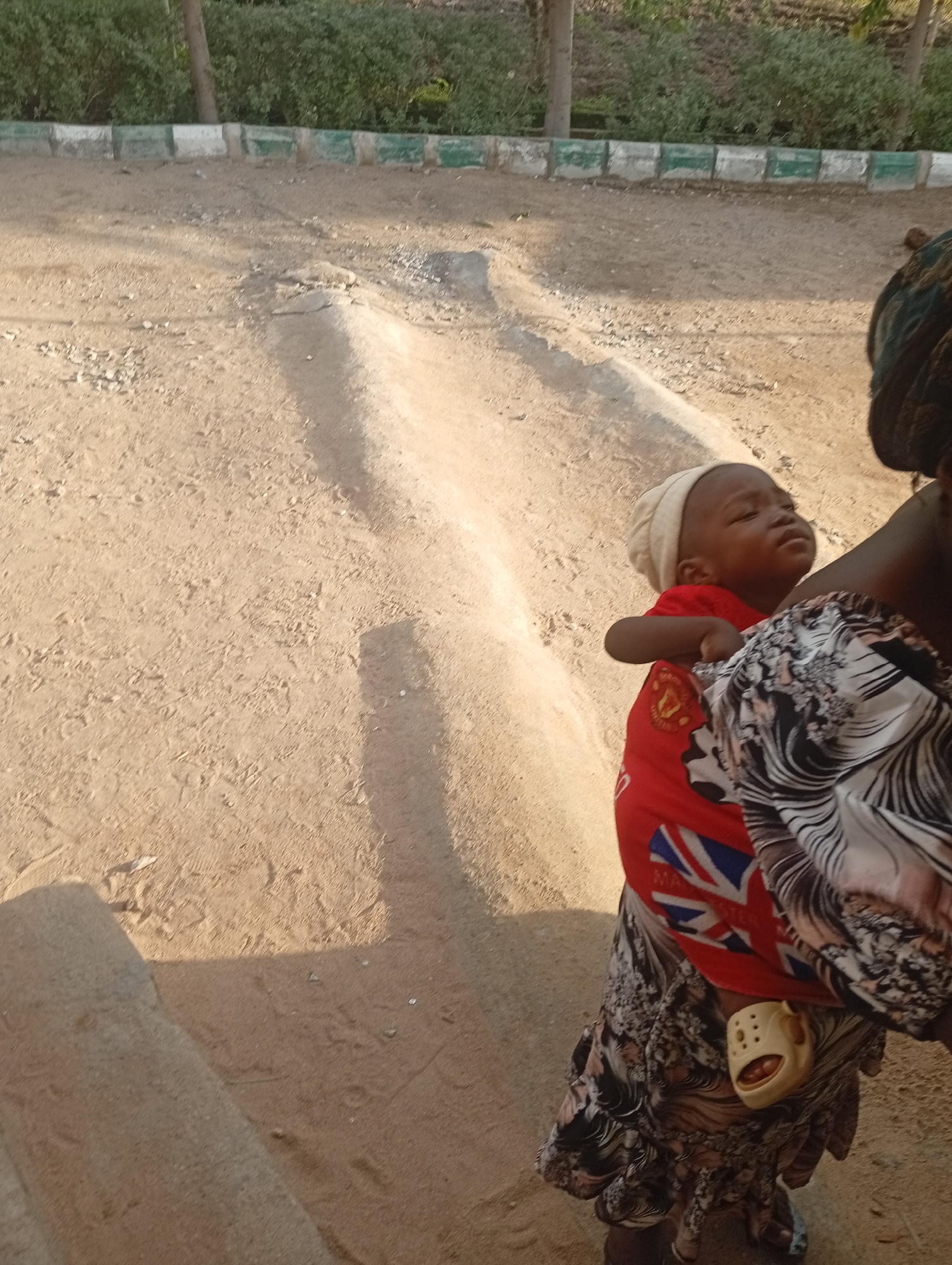 The image shows a close-up of a baby being carried by an adult. The baby is wearing a light-colored cap and a red t-shirt with a Union Jack design. The adult is wearing a patterned wrap and a dark headscarf. The baby is facing upwards and appears to be asleep or looking upwards.
The scene is outdoors, with a dirt path in the background. There is some greenery and a low wall with alternating white and green sections along the top of the path. A shadow falls across the path, suggesting it is daytime, likely late afternoon or early morning due to the angle. The lighting is bright and warm, indicating sunny weather. The overall setting appears to be a somewhat rural or undeveloped area. A single light-colored shoe with holes is attached to the adult's wrap. The textile of the wrap is detailed with a black and white swirly pattern. The baby's t-shirt has a visible text with red and white logos and writing.