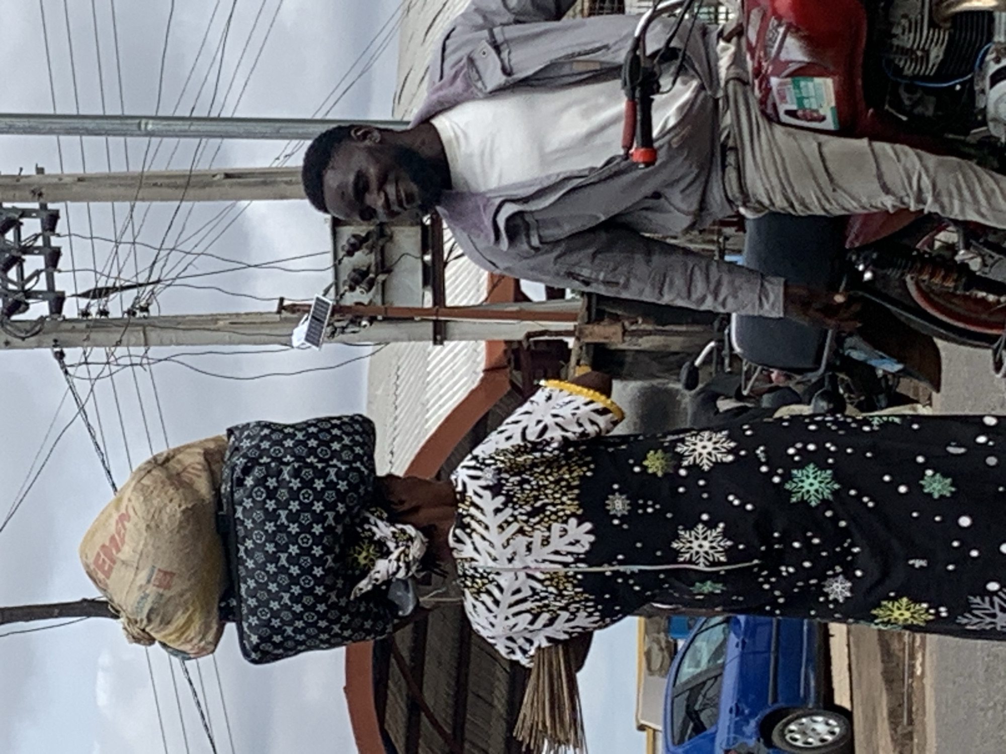 This image depicts an outdoor street scene in Shendam, Nigeria, under an overcast sky.
In the left foreground, a woman is observed from behind, carrying a substantial load balanced on her head. The load comprises a dark-colored, rectangular bag patterned with light-colored stars and symbols, surmounted by a light brown sack prominently labeled "CEMENT." She wears a long, dark-colored dress adorned with white and yellow snowflake-like patterns on the upper half and smaller, multicolored snowflake patterns with white dots on the lower half. A bundle of brooms is held in her left hand, extending horizontally.
In the right foreground, a man is seated on a red motorcycle with a black seat. He is facing partially towards the woman, exhibiting a mild smile or pleasant expression. He has short dark hair and a beard, and is dressed in a white t-shirt, a grey zippered jacket with a visible purplish collar lining, and light-colored trousers.
The background features several utility poles supporting a complex network of electrical wires against the grey sky. A small solar panel device with an attached light is mounted on one of the poles. To the far left, a blue car, identified as a Peugeot 206, is partially visible. Sections of buildings with corrugated metal roofs are also discernible in the mid-ground. The ground surface appears to be a paved road or an unpaved path.