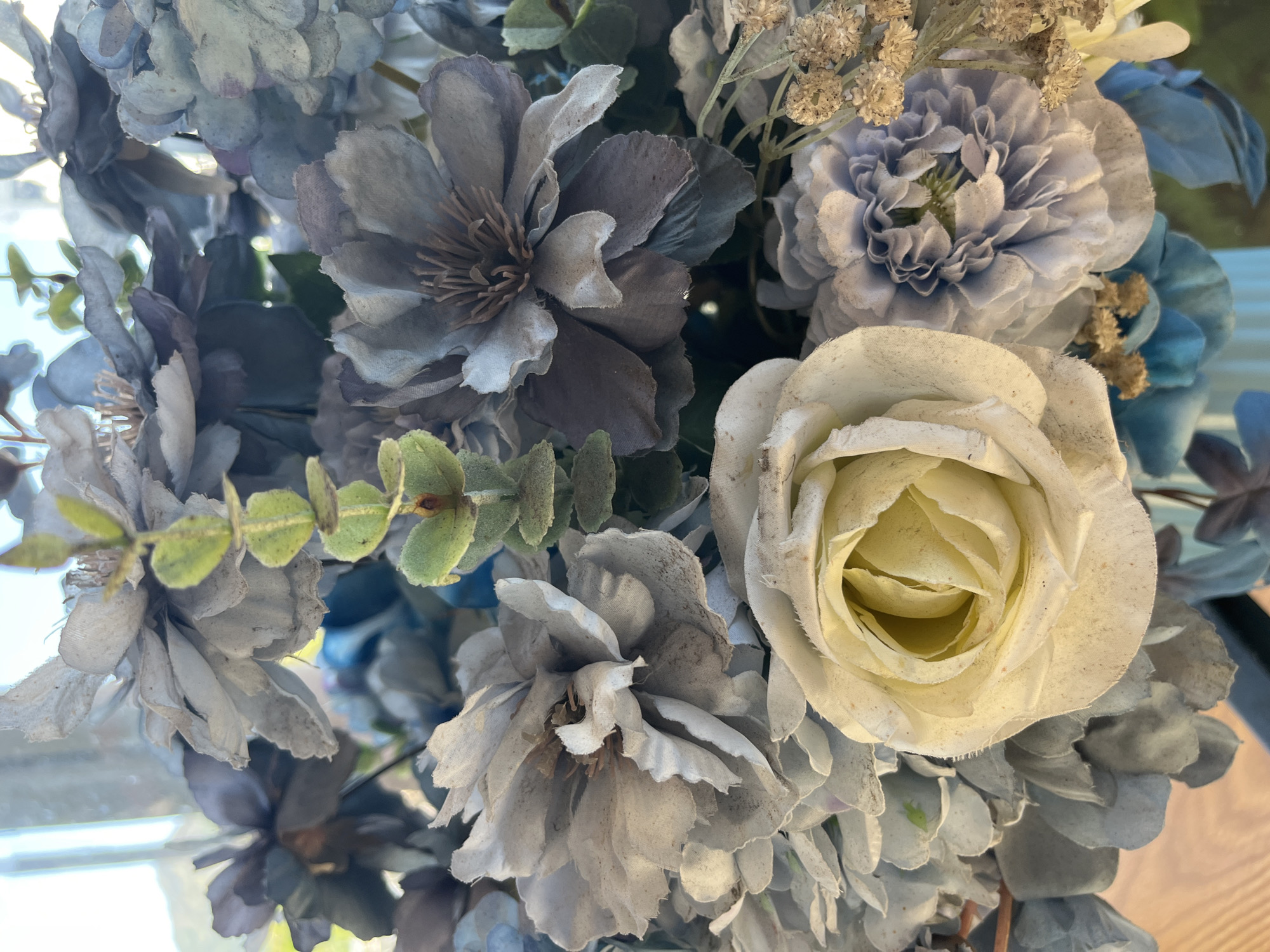 The image shows a close-up of a bouquet of artificial flowers. The flowers are predominantly in shades of blue, gray, and white. The bouquet includes a white rose in the foreground, surrounded by other flower types with varying shapes and textures. There are some green leaves interspersed among the flowers. The scene appears to be indoors, but no background details are visible to establish a specific location.