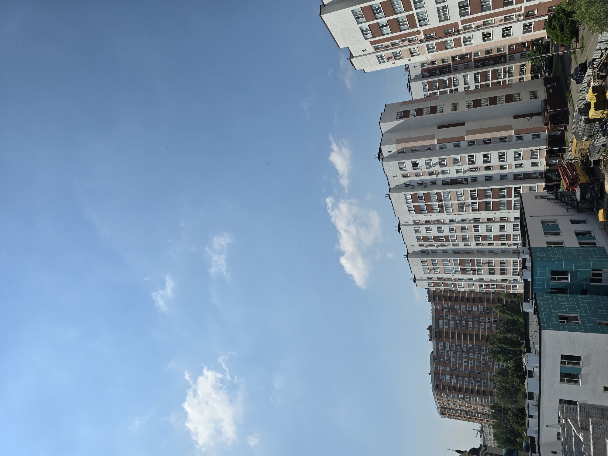 This is a high-angle, wide shot of a residential area in Sofiivska Borshchahivka, Ukraine, on a clear, sunny day. The sky is a vibrant blue with scattered white clouds. In the foreground, a low-rise building with white and teal facades is visible, with visible construction materials and equipment like a crane, trucks, and stacks of supplies. Behind this, several tall, multi-story apartment buildings dominate the scene. The apartment buildings are modern, with white and reddish-brown facades, numerous windows, and balconies. One large building in the background has a distinct curved, brown facade. Trees and greenery are present between the buildings, suggesting a developed urban landscape. There are no people or animals visible in the image. The overall impression is one of urban development and ongoing construction.