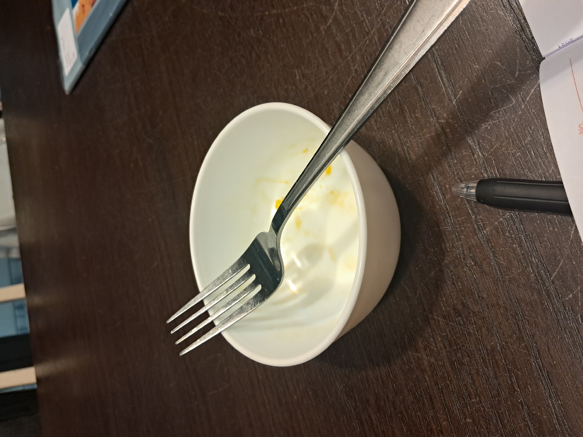 A fork rests across a white bowl on a dark wooden table. The bowl appears to have remnants of food, possibly eggs or a creamy sauce, with yellow and white residue visible on the interior. The fork is metallic, with four tines, and is positioned diagonally, with the tines entering the bowl. To the lower right of the bowl, a black pen is partially visible, and a corner of a notebook with the year "2021" printed on it can be seen. The background is blurred, but suggests an indoor setting with furniture. The location of Bareng Lor, Indonesia, is not discernible from the image itself.