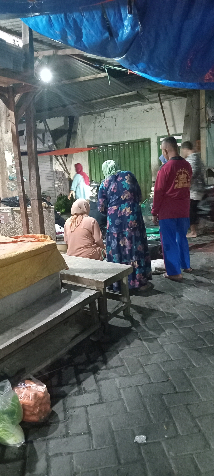 In Babat, Indonesia, at night, four individuals are visible within a marketplace. A man wearing a red long-sleeved shirt and blue pants stands on the right, facing away from the camera. Next to him, a woman in a dark floral dress and green hijab is also facing away. To her left, a person in a peach-colored headscarf and shirt is seated, facing away from the camera. Further left, a person in a light blue top and red headscarf is visible in the background, also facing away. The scene is set under a corrugated metal roof covered by a blue tarp. Wooden beams and posts support the structure. A light illuminates the area from above. In the foreground, to the left, are two bags of vegetables. A wooden bench and a table with a yellow covering are also present. The ground is paved with grey interlocking bricks, and shadows are cast by the objects and people.