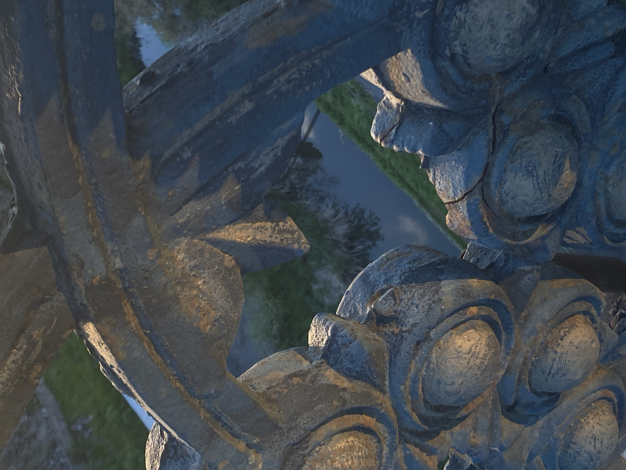 A close-up, angled shot shows part of a decorative metal structure, possibly a bridge or railing, with an ornate, circular design. The metal is weathered and has a blue-grey hue. Through openings in the metalwork, there is a view of a green, grassy area and a narrow, dark body of water or road. The image appears to have been taken during the day.