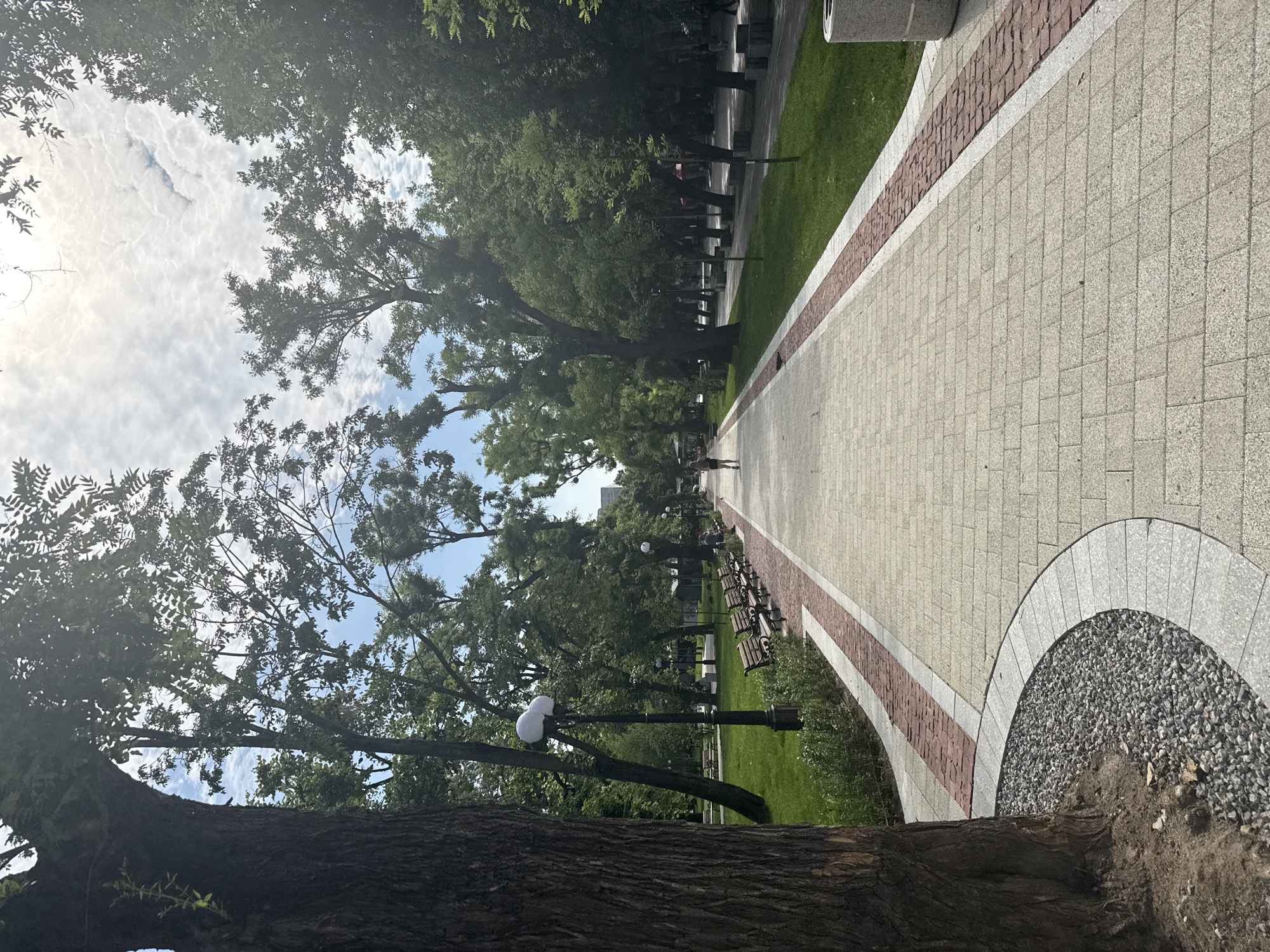 The image depicts a pedestrian walkway in a park, likely in the city of Stara Zagora, Bulgaria. The walkway is paved with light-colored rectangular stones and bordered by a strip of red brick pavers and a strip of white concrete. Lush green grass flanks the walkway on either side, dotted with trees and park benches. Several streetlights with dual spherical lamps are visible along the path. In the foreground, on the left, a large tree trunk with rough bark dominates the scene, surrounded by small grey gravel. In the background, a lone figure, appearing to be a woman with dark hair wearing a dark top and light shorts, walks away from the camera down the path. Other figures are visible further in the distance, suggesting the park is open to the public. The sky above is partially obscured by the canopy of trees and is filled with scattered white clouds against a bright sky. The scene appears to be set during the daytime, with ample natural light.