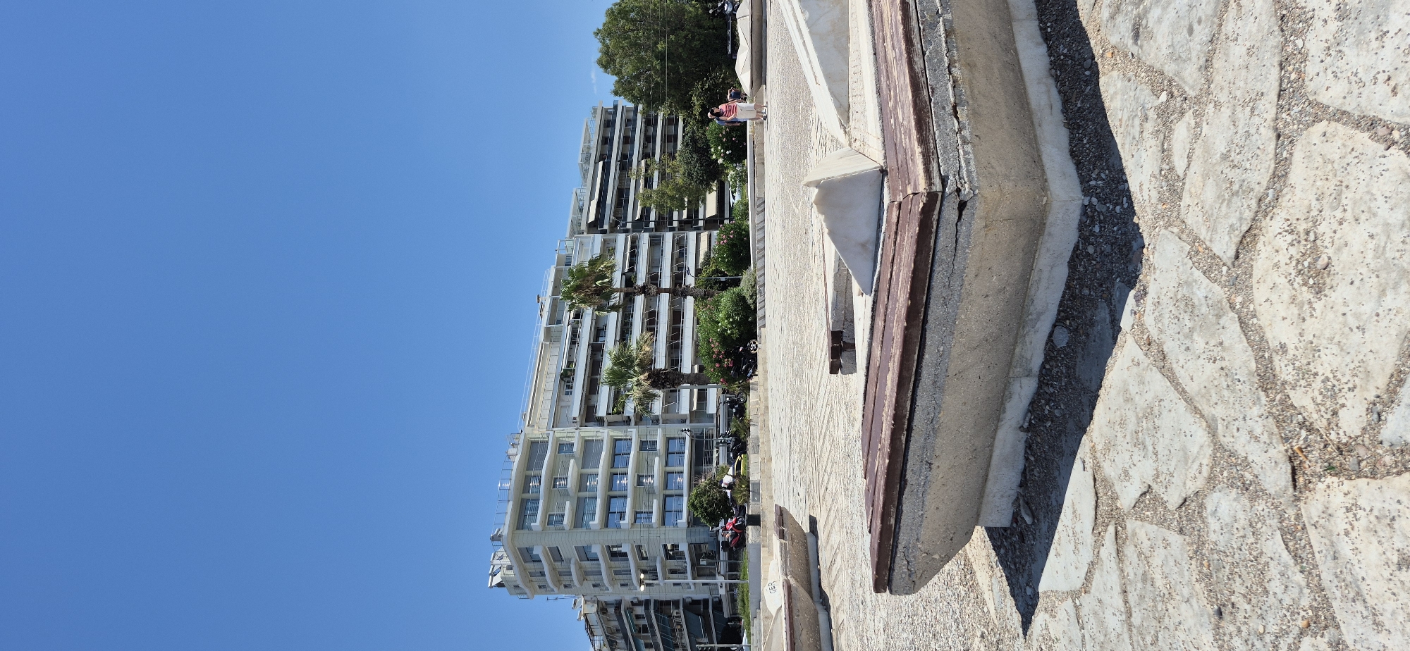 The image depicts a sunny day in Paleo Faliro, Greece. In the foreground, a concrete bench with wooden slats is prominently featured, situated on a paved area with irregular stone slabs and gravel. The bench is weathered, with visible cracks in the concrete. In the middle ground, two people, a woman and a man, are walking on the paved area. The woman is wearing a pink top and white shorts, while the man is in a light-colored shirt and shorts. Behind them, a line of palm trees and other vegetation separates the paved area from a multi-story white building with numerous balconies and windows. Further in the background, other buildings and trees are visible. The sky above is a clear, bright blue. The scene suggests a public outdoor space, possibly a promenade or a park area along the coast.