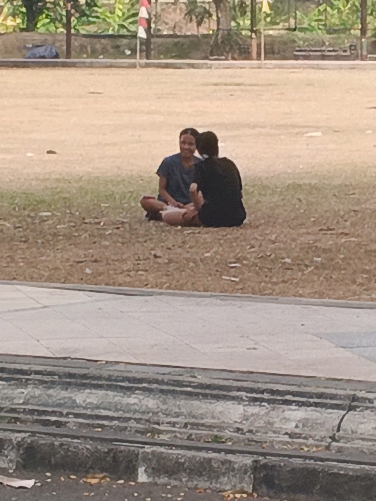 Two individuals are seated on a grassy field. The person on the left is wearing a dark blue t-shirt and appears to be smiling. The person on the right is wearing a black t-shirt. They are positioned facing each other, with their legs crossed. The environment is an open area with dry grass and some fallen leaves. In the background, there is a line of trees and foliage, as well as a fence and a pole with a red and white flag, suggesting a public park or recreational area in Kalirejo, Indonesia. A paved walkway is visible in the foreground.