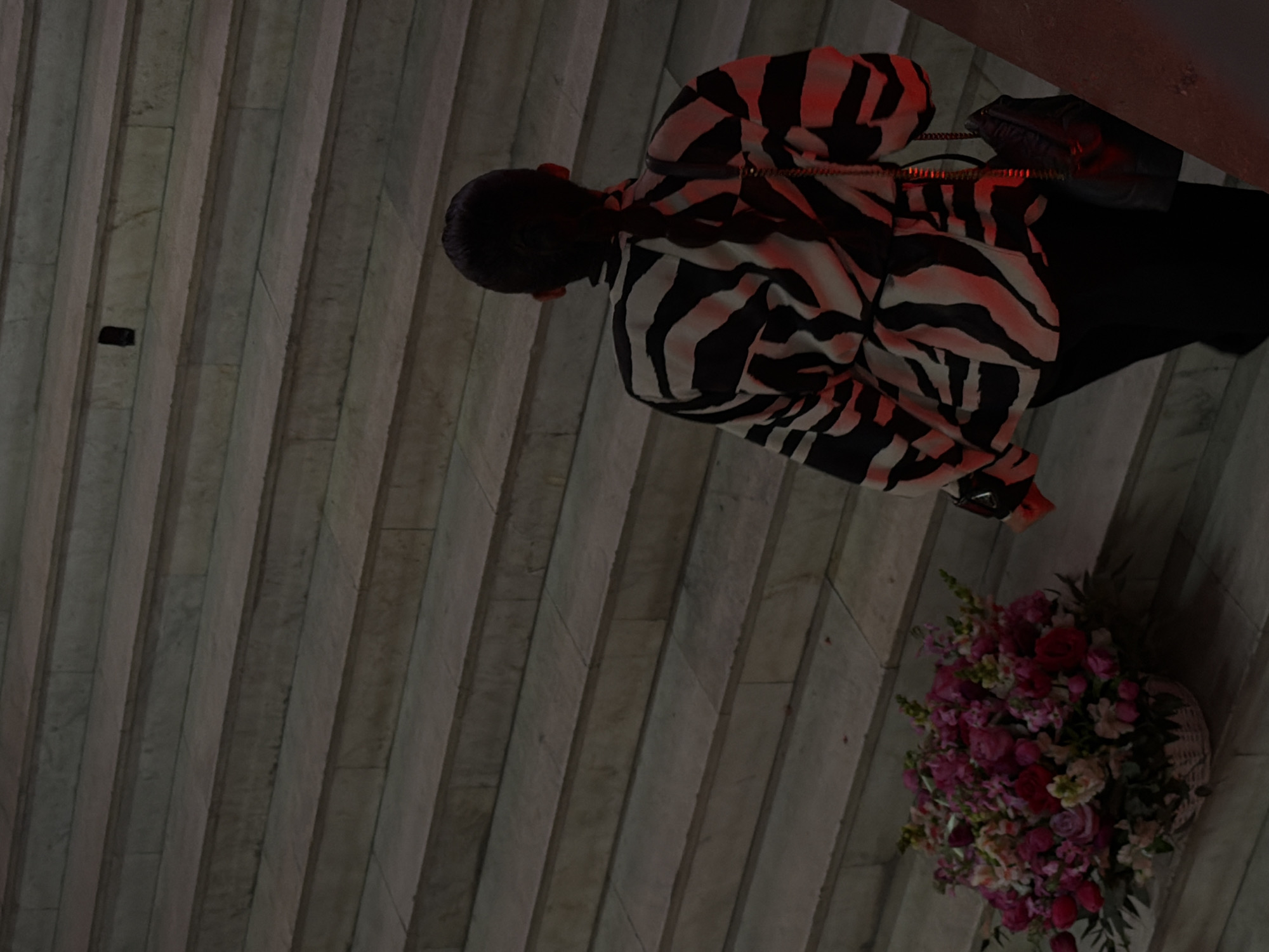A high-angle, softly lit photograph shows a person standing on a pale gray, ridged stone surface in Yerevan, Armenia. The person is wearing a black-and-white zebra-striped jacket and black pants, and has a small black shoulder bag. A large bouquet of pink, red, and white flowers sits near the person's feet on the stone surface. The location appears to be an outdoor public space, likely a walkway or plaza. The lighting is somewhat dim and uniform, casting soft shadows. A small, dark object is visible in the background on the stone surface.