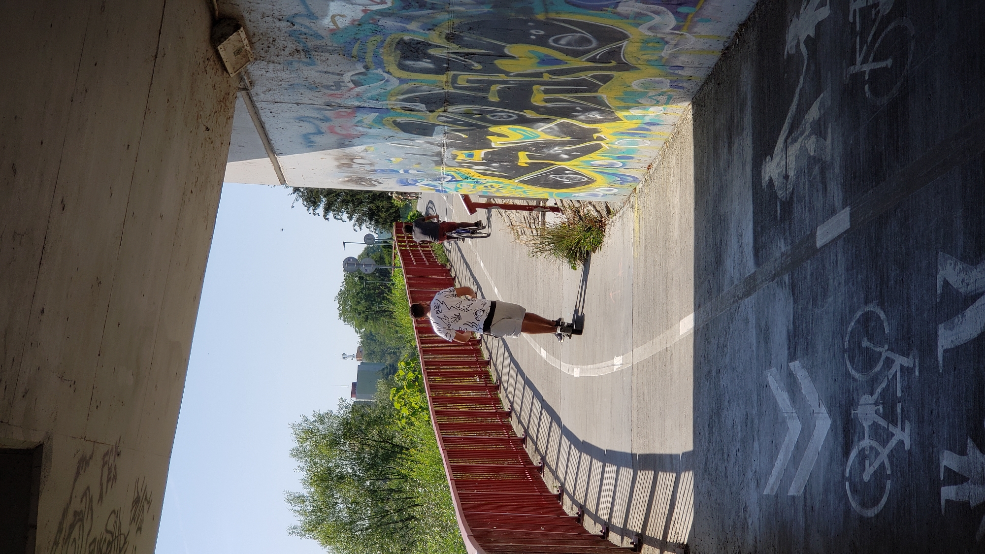 The image shows a pedestrian and bicycle path under a concrete overpass. The path is marked with white lines and painted symbols of bicycles and pedestrians. On the right side of the path, there is a concrete wall covered in colorful graffiti. On the left side, there is a red railing and lush greenery.
In the foreground, a man is riding an electric scooter, wearing a white t-shirt with a black pattern and grey shorts. Behind him, another person is riding a bicycle. The path curves to the left, and in the distance, more people are seen on bicycles. The sky is clear and blue.
The scene is located on the path under the bridge leading to the Sibírska housing estate in Prešov, Slovakia. This path is known for its bike lane and pedestrian walkways, separated by a red railing. The graffiti on the wall is a common feature of this area, adding a touch of urban art to the environment. The overpass provides shade on a sunny day, creating a contrast between light and shadow on the path.
