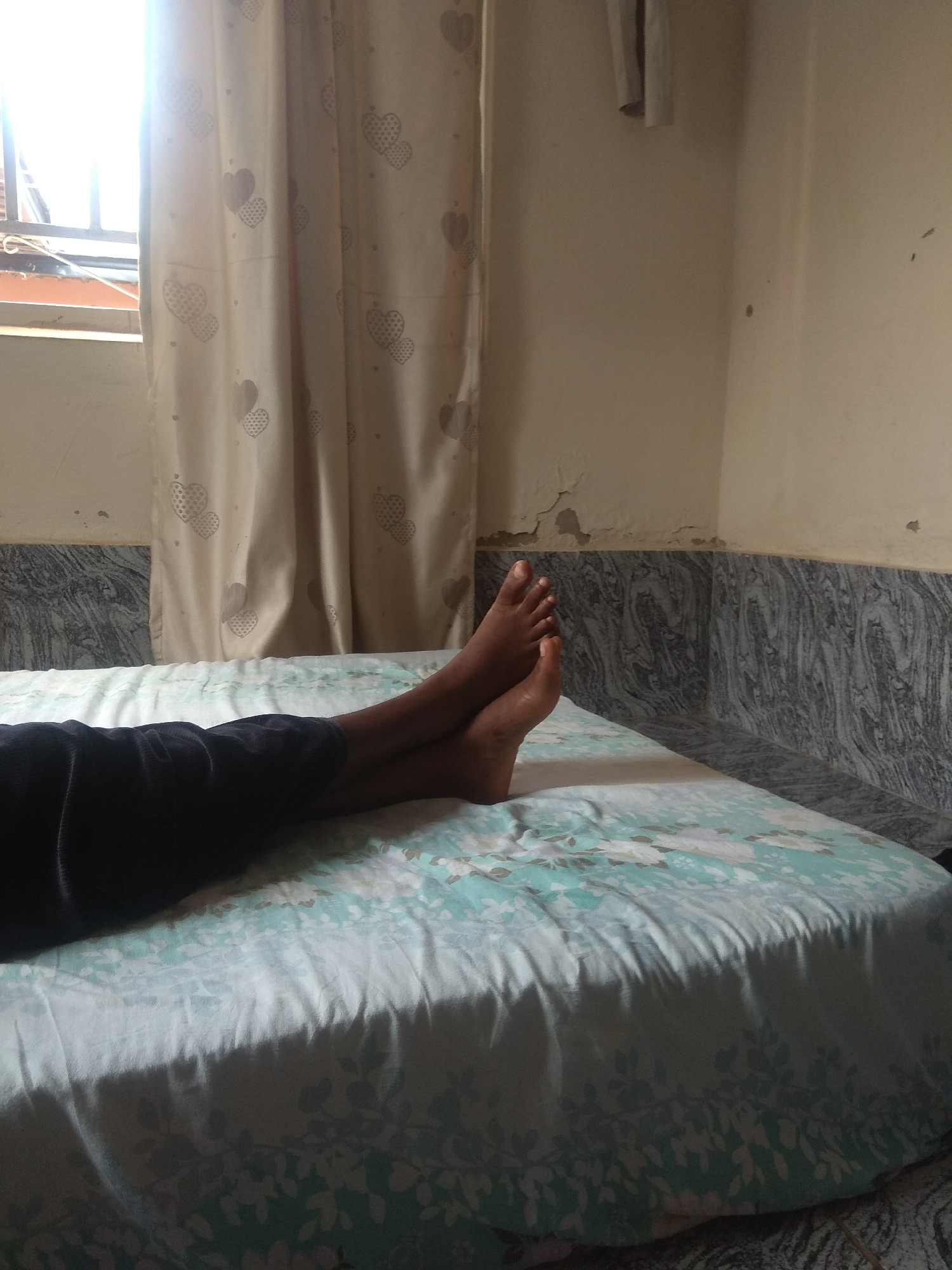 A person's legs and feet are visible, crossed at the ankles, resting on a bed covered with a floral patterned sheet. The person is wearing dark-colored shorts. The bed is positioned in a room with beige walls and a marble-patterned baseboard. A light-colored curtain with a heart pattern hangs behind the bed. Sunlight streams in from a window to the left of the frame. The scene suggests a relaxed or resting posture within a domestic setting.