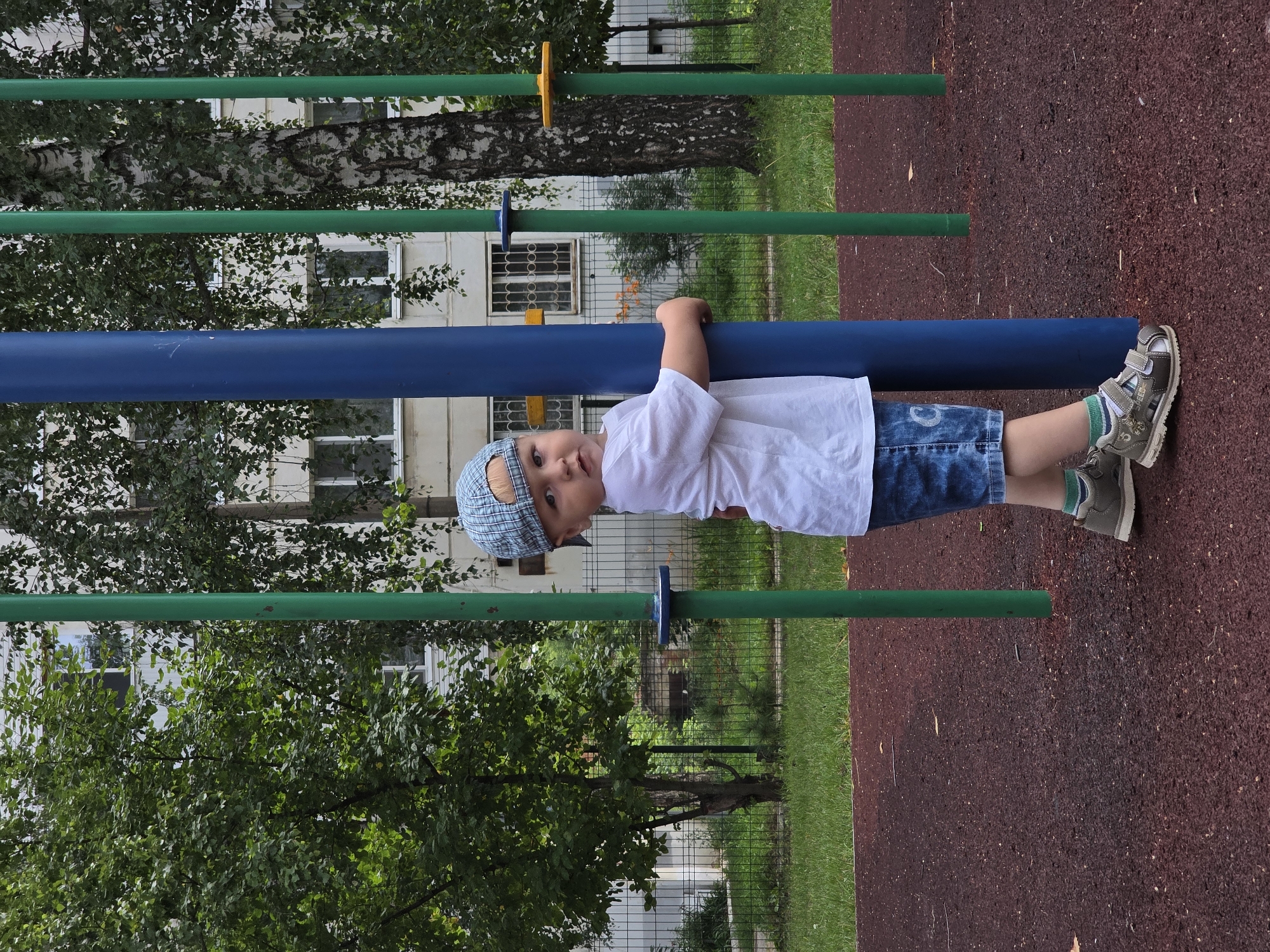 A young boy, wearing a white t-shirt, denim shorts, a checkered blue baseball cap, and grey sandals with green and white striped socks, stands on a red rubberized playground surface, clutching a thick blue vertical pole. He is facing the camera with a slight upward tilt of his head and a curious expression. To his right, several green vertical poles and a birch tree with dark bark and light markings are visible. Behind the playground equipment, a multi-story building with white siding and numerous windows is partially obscured by foliage from trees, some with hanging branches. A green metal fence runs horizontally across the middle ground. The overall scene is an outdoor urban playground environment. The location is a playground in the city of Chaikovsky, Russia.