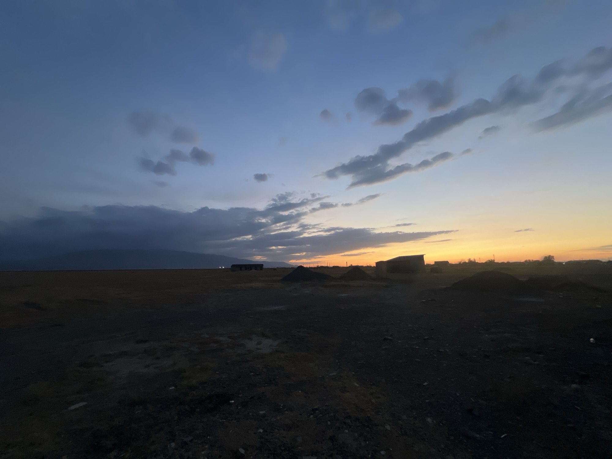 The image captures a desolate landscape under a twilight sky, located in the vicinity of Ақши, Kazakhstan. The sky transitions from a deep blue at the top to a warm orange glow near the horizon, indicative of either sunrise or sunset. Scattered clouds, varying in color from dark gray to light gray and white, dot the sky.
The foreground is a barren, dark, and uneven terrain with scattered small rocks and debris. In the middle ground, several low-lying structures, possibly dilapidated buildings or sheds, are silhouetted against the sky. Piles of dark material, resembling gravel or coal, are also visible. A distant mountain range is faintly discernible on the left side of the image, shrouded in a darker, atmospheric haze. On the far right, faint lights suggest the presence of further structures or human activity. No people are visible in the frame. The overall scene conveys a sense of remoteness and an undeveloped or industrial environment.