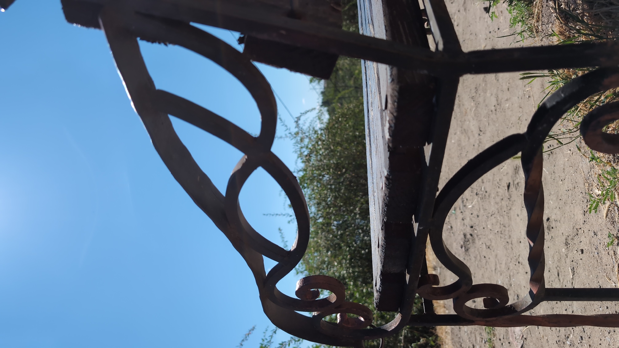 A close-up, low-angle shot shows the armrest and seat of a rustic metal bench on a sunny day. The bench has a dark, weathered metal frame with decorative scrollwork and a twisted metal element. The seat appears to be made of wooden planks, also showing signs of age and weathering. In the background, out-of-focus green foliage and a clear blue sky are visible. The ground beneath the bench is sandy and dry, with sparse dry grass. The sunlight casts highlights on the metalwork. The image is taken outdoors, likely in a park or garden setting. The location in Mykolaiv, Ukraine is not specified or discernible from the image.
