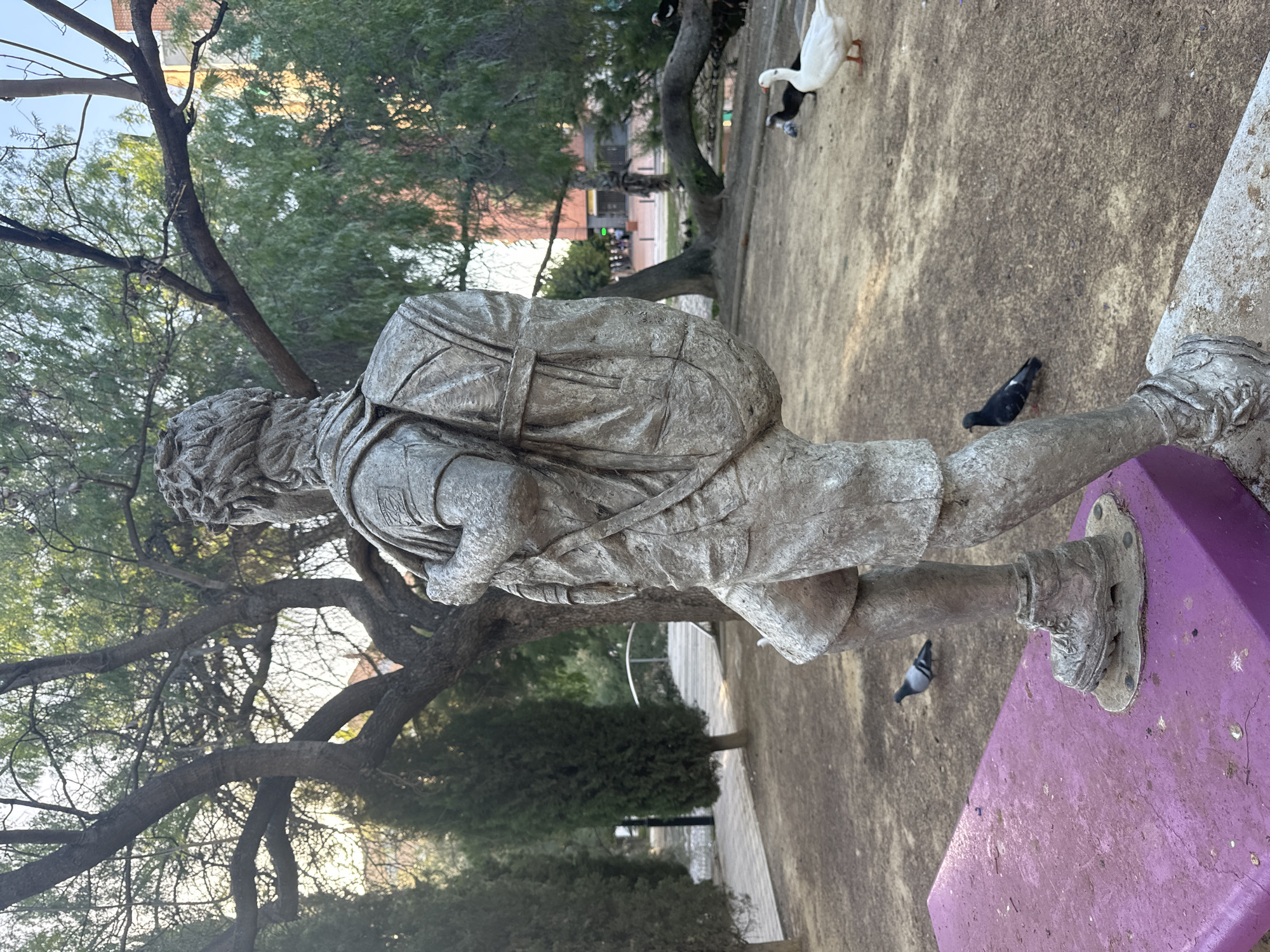 A statue of a person wearing a backpack and shorts stands in a park, with pigeons and ducks visible. The statue is made of a weathered metallic material and is positioned on a purple platform. The background features trees, a bridge, and buildings in the distance. The scene appears to be in an urban park setting, likely in the city of Murcia, Spain, given the architectural style of the distant buildings and the presence of Mediterranean vegetation. The lighting suggests it is either early morning or late afternoon.