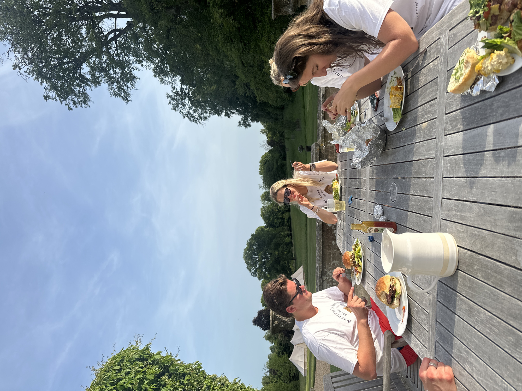 Three individuals are seated at a wooden table outdoors, engaged in a meal. A woman with long brown hair is observed eating from a plate, another woman with blonde hair is seated at the table, also engaged in eating. A man with dark hair wearing sunglasses is seated at the table, also engaged in eating. Food is visible on the table, including burgers, salad, and corn. A white pitcher and a bottle are also present on the table. The surroundings include trees, sky, and grass. In the background is a light-colored stone wall. The location appears to be a garden or outdoor eating area.