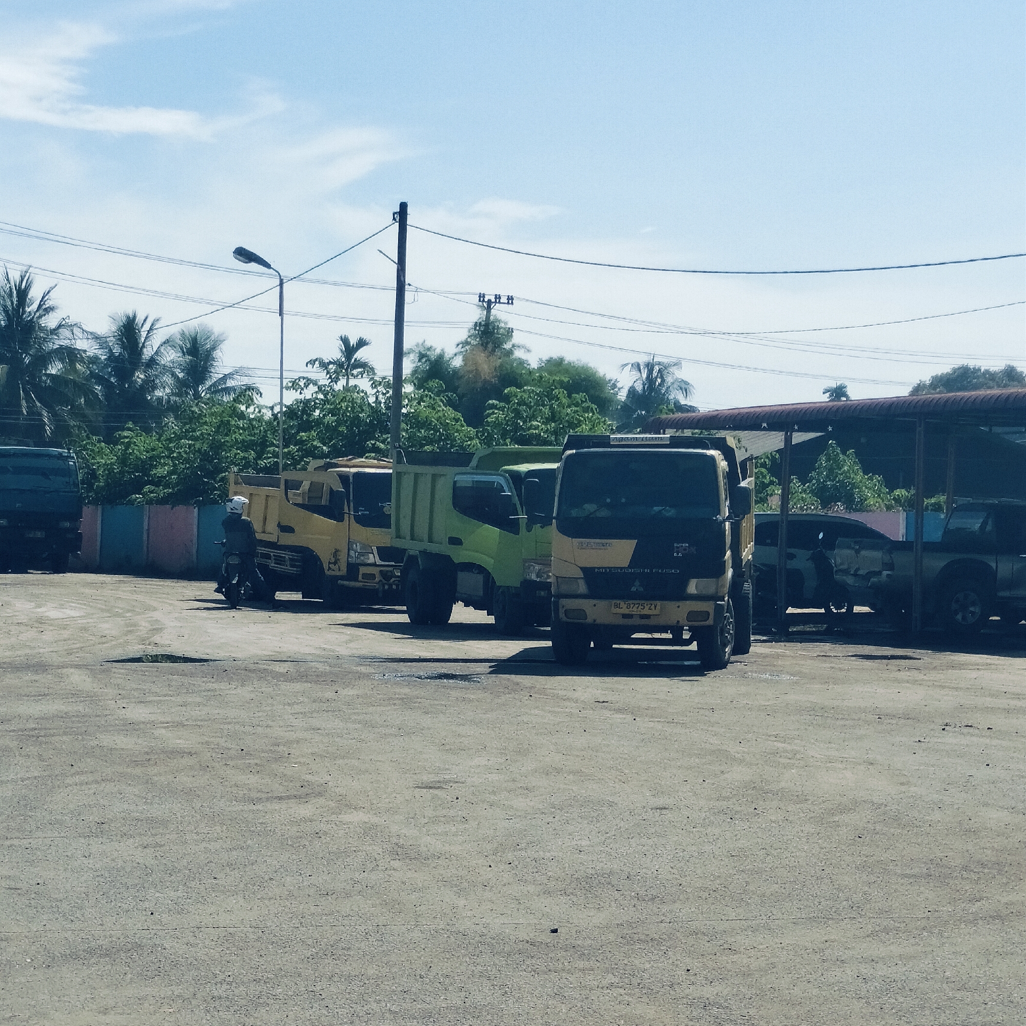 This image captures a bright daytime scene at an outdoor vehicle yard or parking area in Cotpuuk, Indonesia. The sky is a clear blue with sparse white clouds, indicative of sunny weather.
The primary subjects are several commercial dump trucks. On the far left, a dark-colored truck is partially visible. Next to it, a yellow dump truck is parked, with a person wearing a white helmet seated on a dark motorcycle near its front. Further right, a green dump truck is parked. Most prominent in the foreground on the right is a black and yellow Mitsubishi Fuso dump truck, identified by text as "MITSUBISHI FUSO" and "SUPER HDX 6.6," displaying the license plate "BL 8775 ZY." To its right, under a corrugated metal shed, several other vehicles, including what appears to be a pickup truck, are parked.
The ground is a light-colored, rough, and uneven surface, possibly cracked asphalt or packed dirt. The background features lush, dense tropical greenery, including palm trees, and a low wall with blue and pink segments. Power lines stretch across the upper portion of the frame, and a street light pole is visible on the left. The scene suggests a casual parking or waiting area for these commercial vehicles.