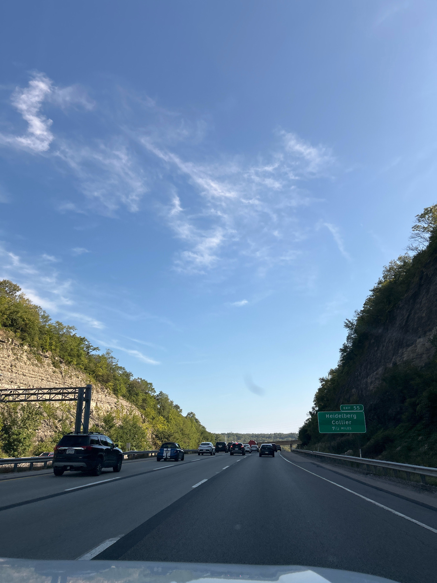The image depicts a scene on a highway in Collier Township, United States of America, on a clear day with scattered clouds. The perspective is from inside a vehicle, looking forward along the multi-lane road. On the right side of the highway, a large green sign indicates "EXIT 55 Heidelberg Collier 1 1/2 MILES". To the right of the sign, a steep, rocky hillside covered with trees rises sharply. On the left side of the highway, a similar, though less steep, rocky hillside is visible, also covered with trees and featuring overhead gantry structures for traffic signs. Several vehicles are on the highway, traveling in the same direction as the viewer. The road surface is asphalt with visible lane markings.