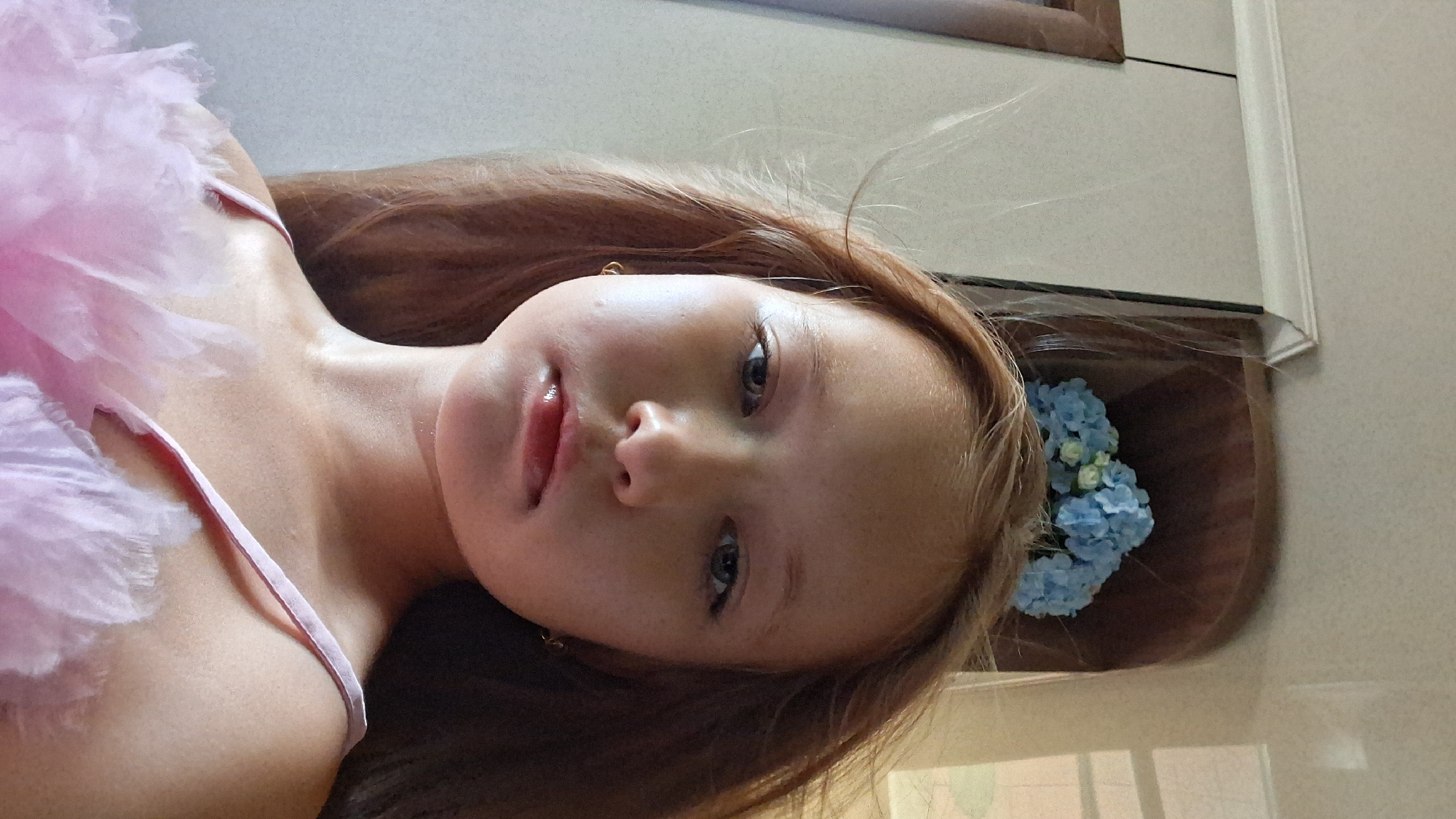 This is a close-up, selfie-style photograph of a young girl, likely a child, lying down and looking upwards towards the camera. She has long, light brown hair that is spread out around her head. Her face is the central focus, with her eyes looking directly at the viewer. She has a slight smile, and her lips are parted. She appears to be wearing a pink, possibly a tulle or feathered garment, on her left side, which covers part of her chest and shoulder. There is a small gold earring visible in her left ear.
In the background, to the right of her head, there is a bouquet of blue flowers, possibly hydrangeas, with some white accents. These flowers are arranged in a container that is mostly out of frame, but appears to be a wooden or dark brown material. Behind the flowers, a reflective surface, likely a mirror or a glossy wall, shows a suggestion of the room. The overall lighting suggests it is daytime, with soft, natural light. The girl's expression is neutral to slightly happy, and her gaze is direct and engaging. There is no visible text in the image. The context provided suggests the location is Semey, Kazakhstan, though there are no specific visual cues within the image to confirm this.